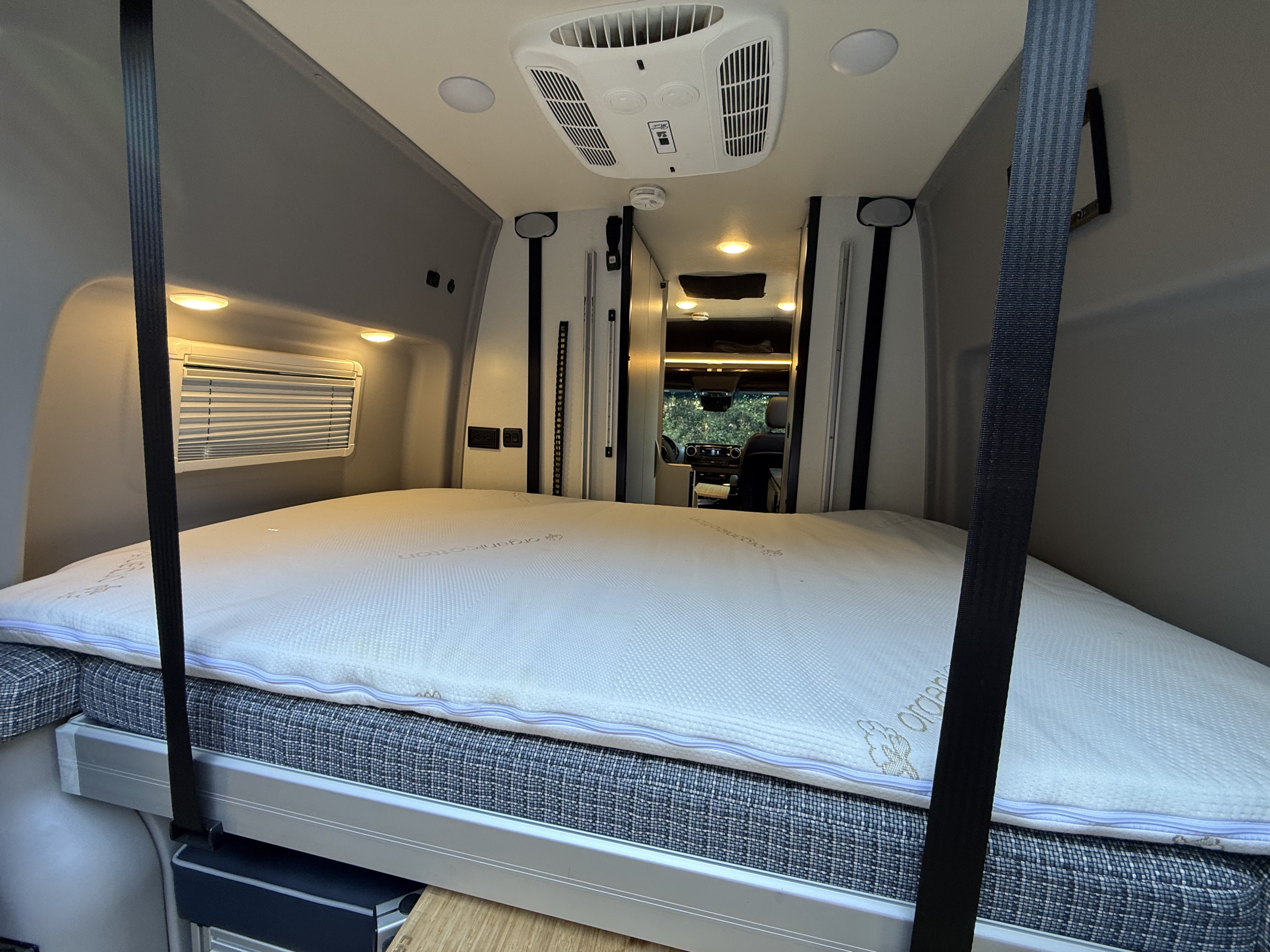 Inside a bright rv rental, a large, comfortable bed with an overhead AC unit and lighting, secured by black straps.. Winnebago Revel 2022
