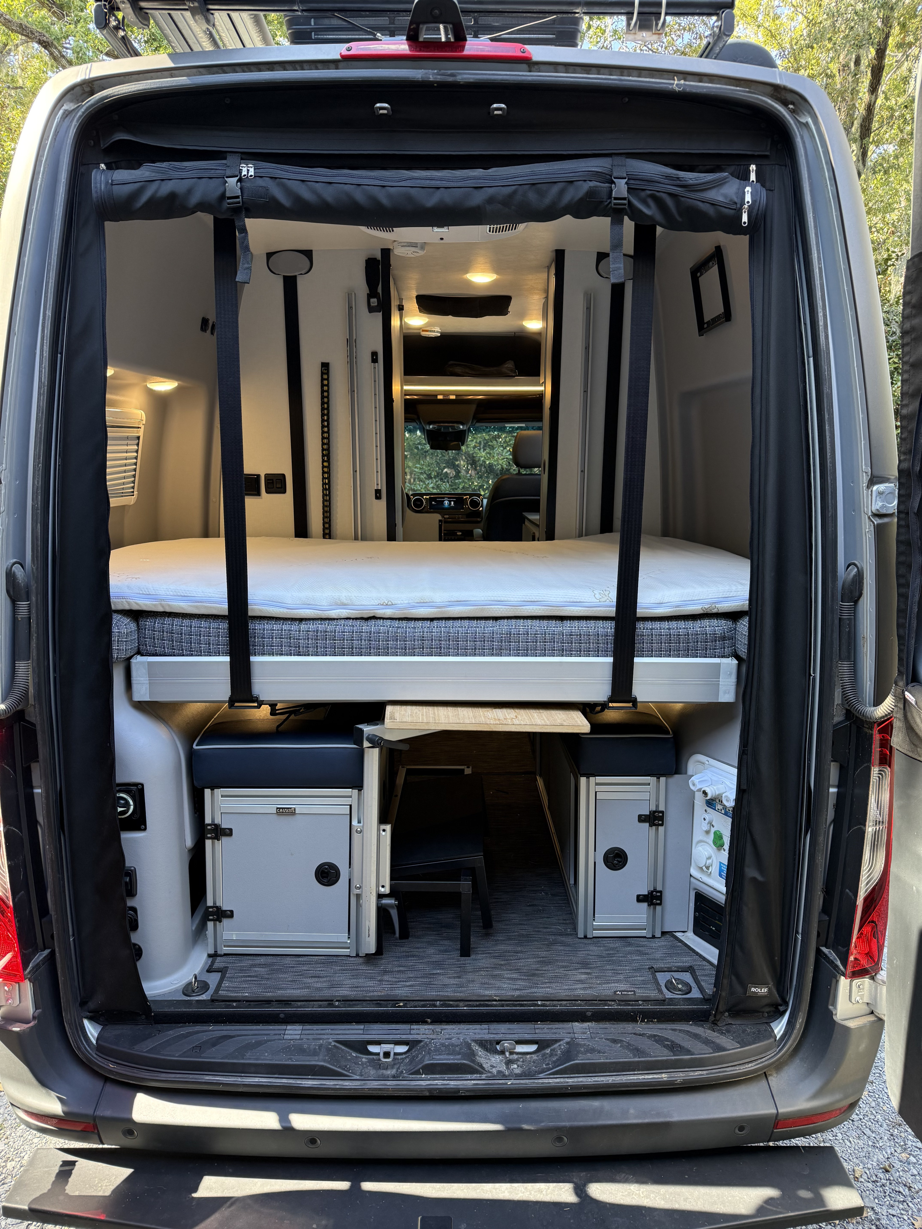 RV rental camper van's rear interior with a raised bed over storage units, a central table, and black privacy curtains.. Winnebago Revel 2022