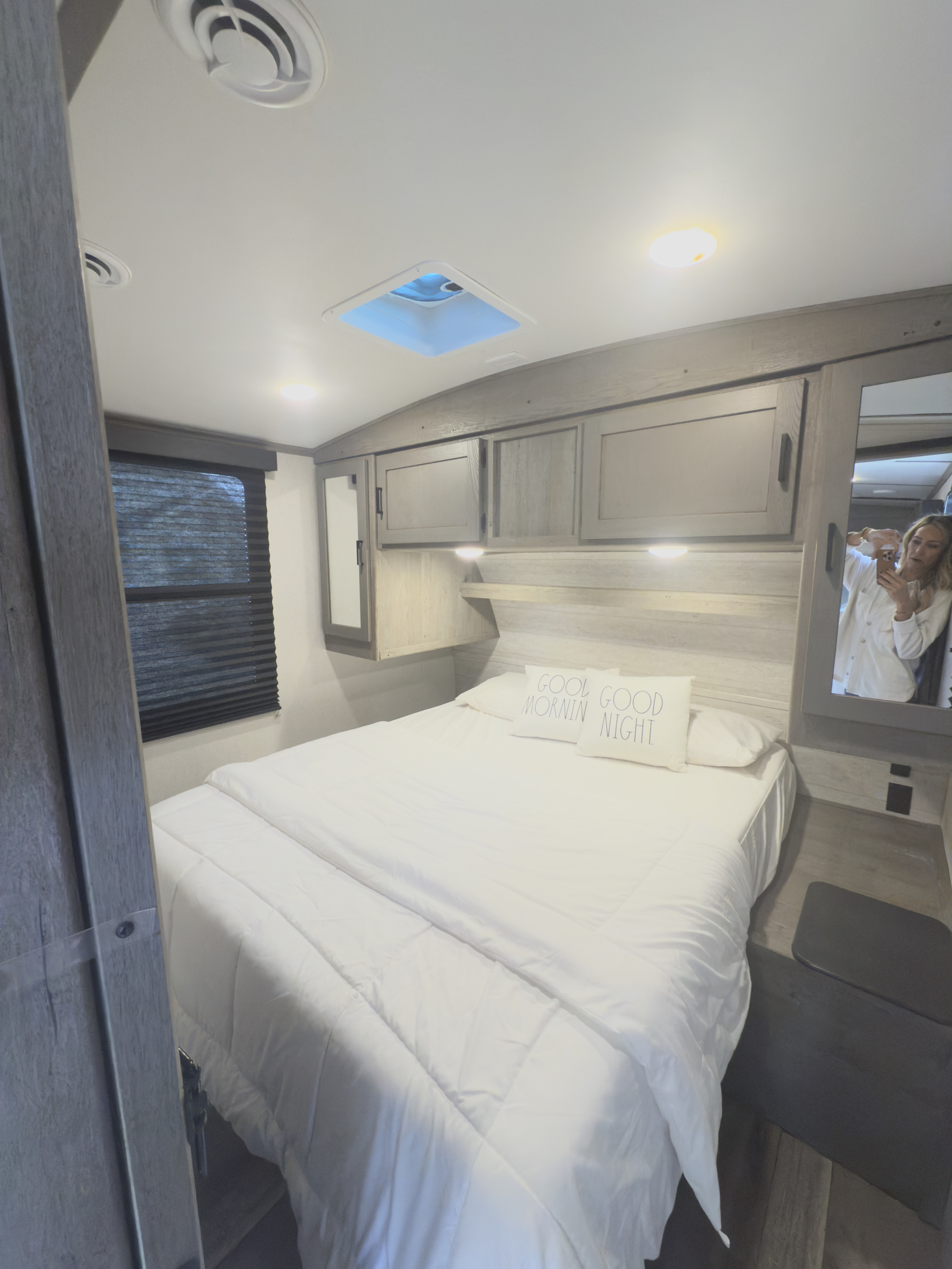 Cozy RV rental bedroom with a white queen bed, overhead cabinets, window, skylight, and mirror (reflection of a person).. Keystone RV Outback Ultra-Lite 2021