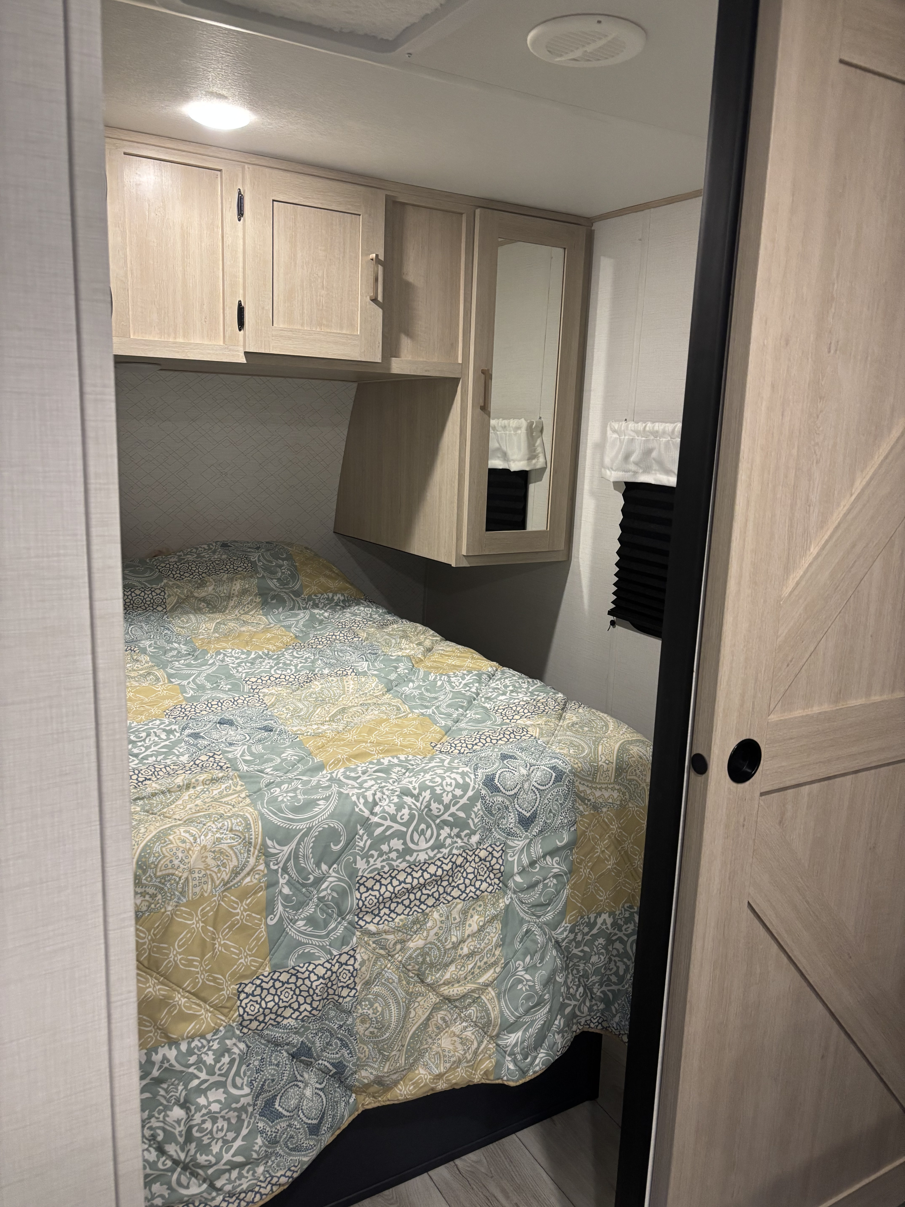 Cozy RV rental bedroom with patterned bed, light wood cabinets, mirrored door, and pleated window blind, offering compact storage.. Forest River, Inc. Primetime Avenger 2025