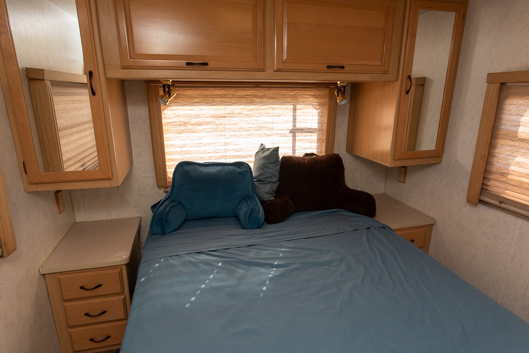 This RV rental bedroom features a comfortable bed with blue and brown pillows, wooden cabinets, and a window with blinds.. Itasca Sundance 1998