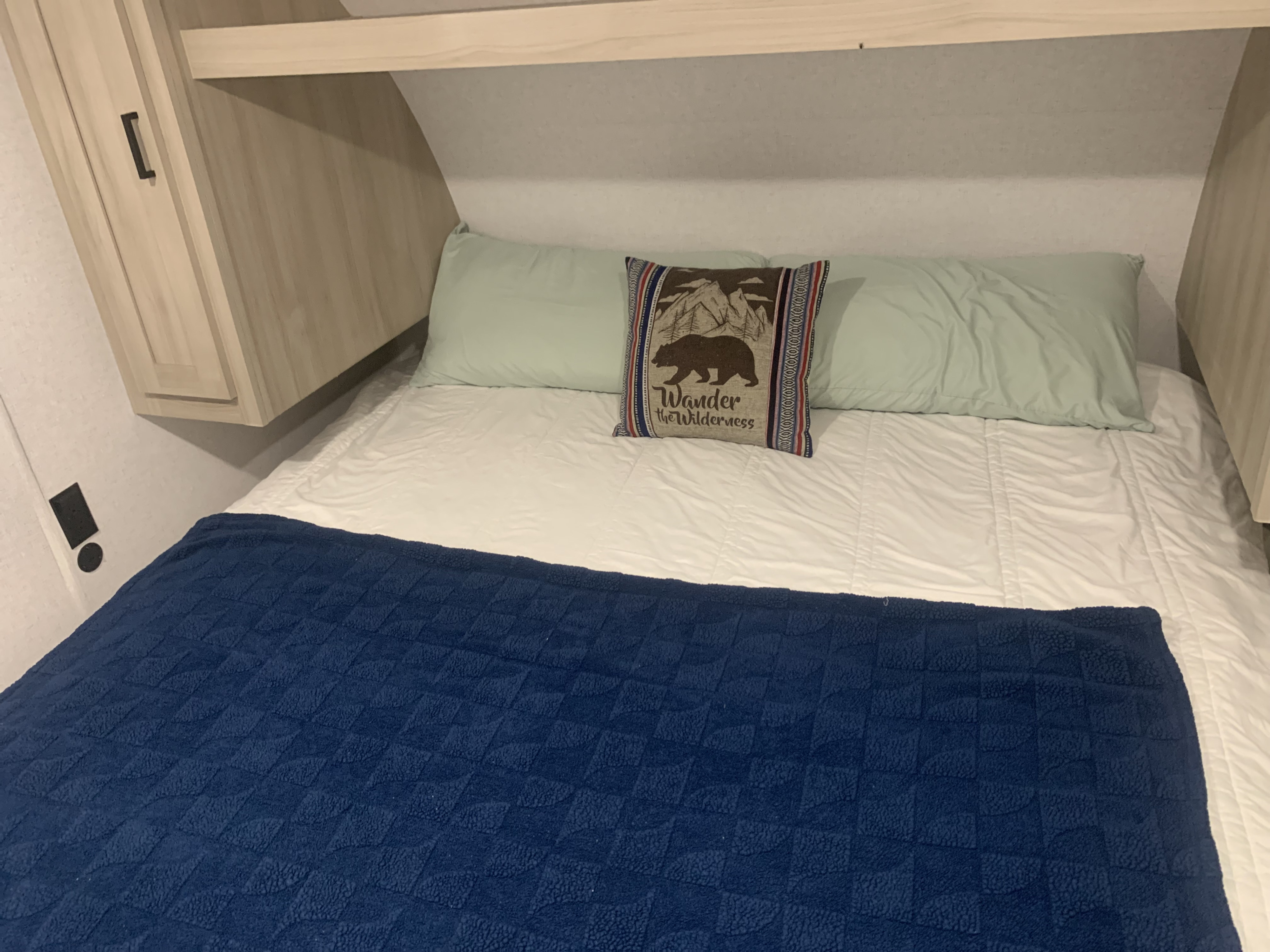 Cozy RV rental bed with light green and decorative pillows, a white comforter, and blue textured blanket, framed by wood cabinetry.. Forest River East to West 2025
