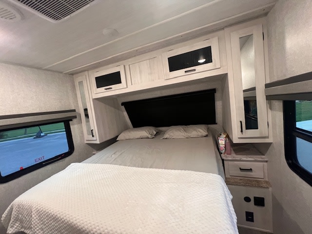 Cozy RV rental bedroom features a comfortable bed with white bedding, overhead cabinets, and convenient side tables with outlets.. East to West Longitude 2024