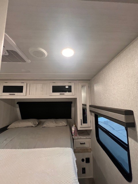 Cozy RV rental bedroom featuring a comfortable bed, overhead cabinets, a small window, and modern interior design.. East to West Longitude 2024
