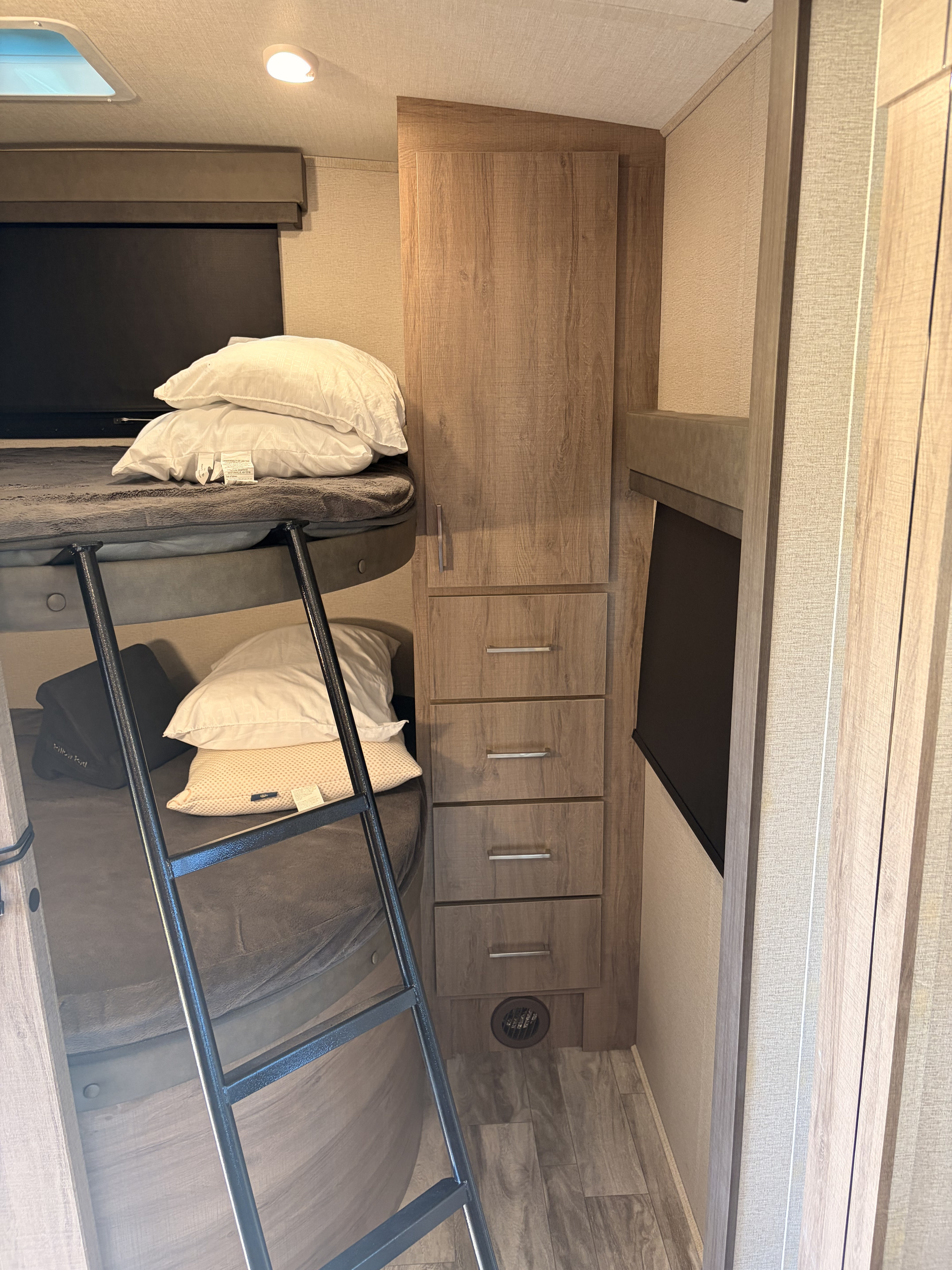 Inside an RV rental: bunk beds with a ladder, adjacent wooden wardrobe with drawers, and a window with a dark blind. Skylight overhead.. Grand Design Imagine 2023