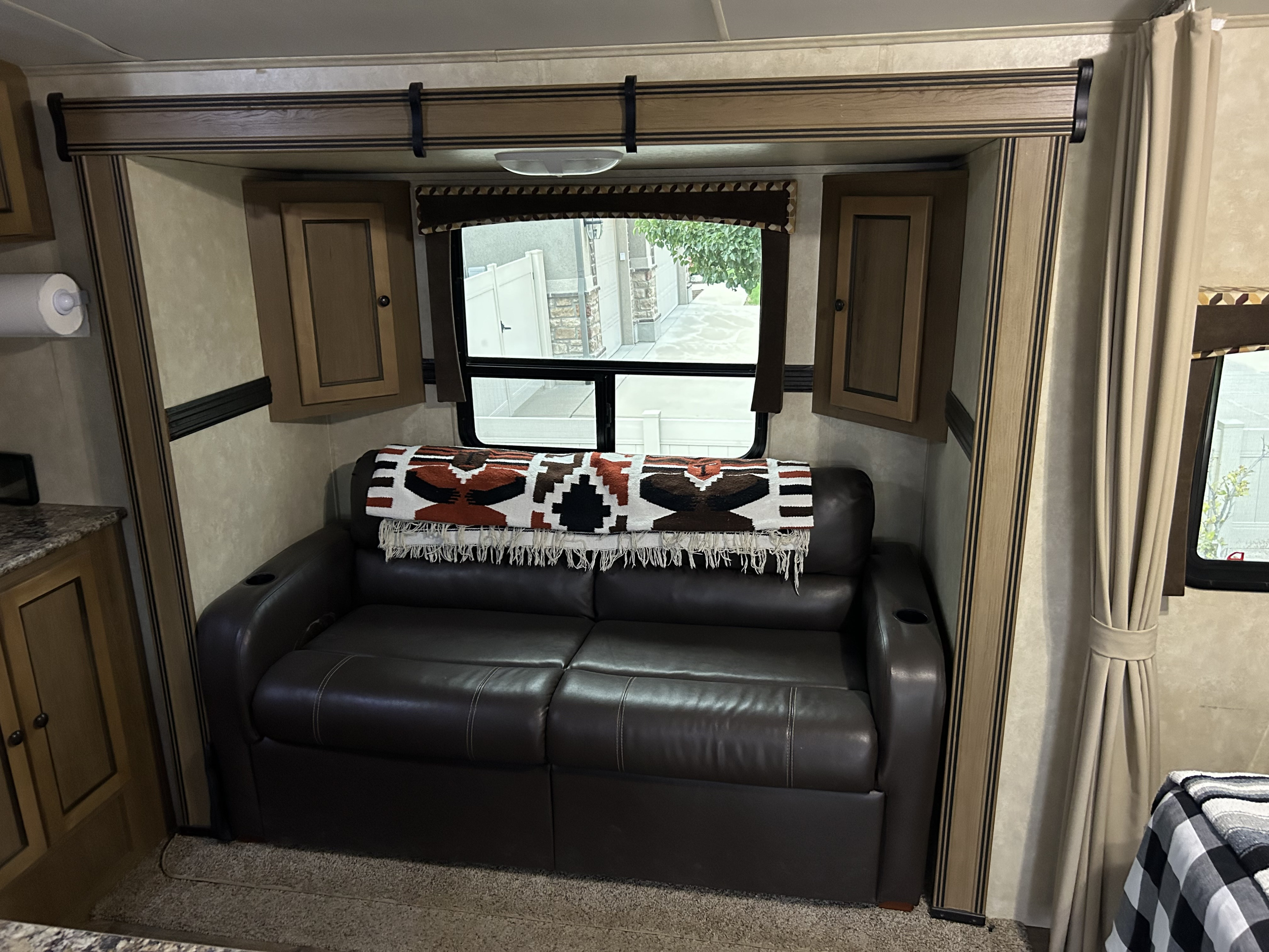 Step into comfort! This RV rental features a dark brown leather sofa with a patterned blanket, window view, and wooden cabinets.. Cruiser RV Shadow Cruiser 2015