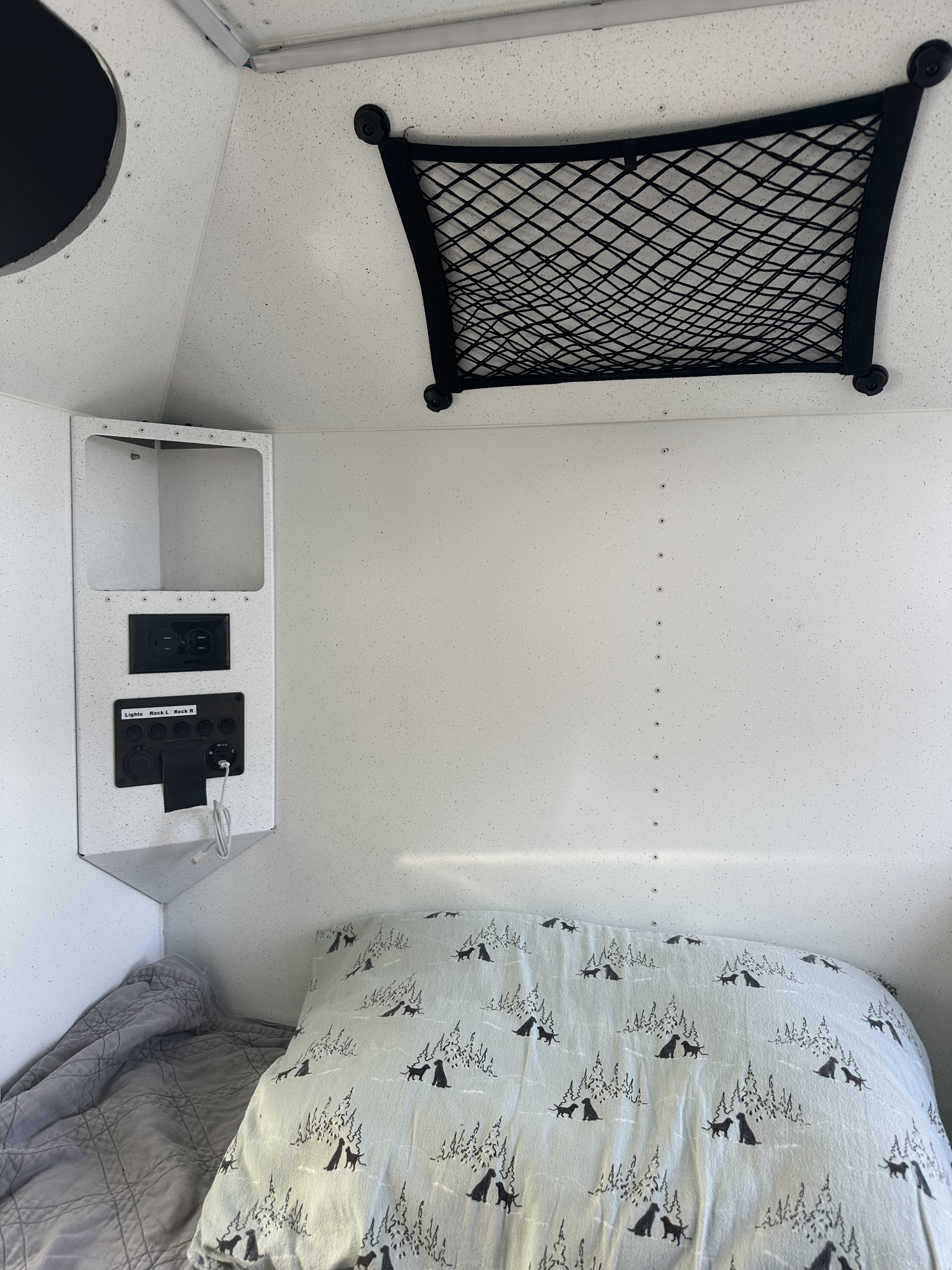 RV rental bed with dog-patterned pillow, power panel (AC/USB outlets), and overhead storage net. Camper interior.. Mammoth Overland HV 2022