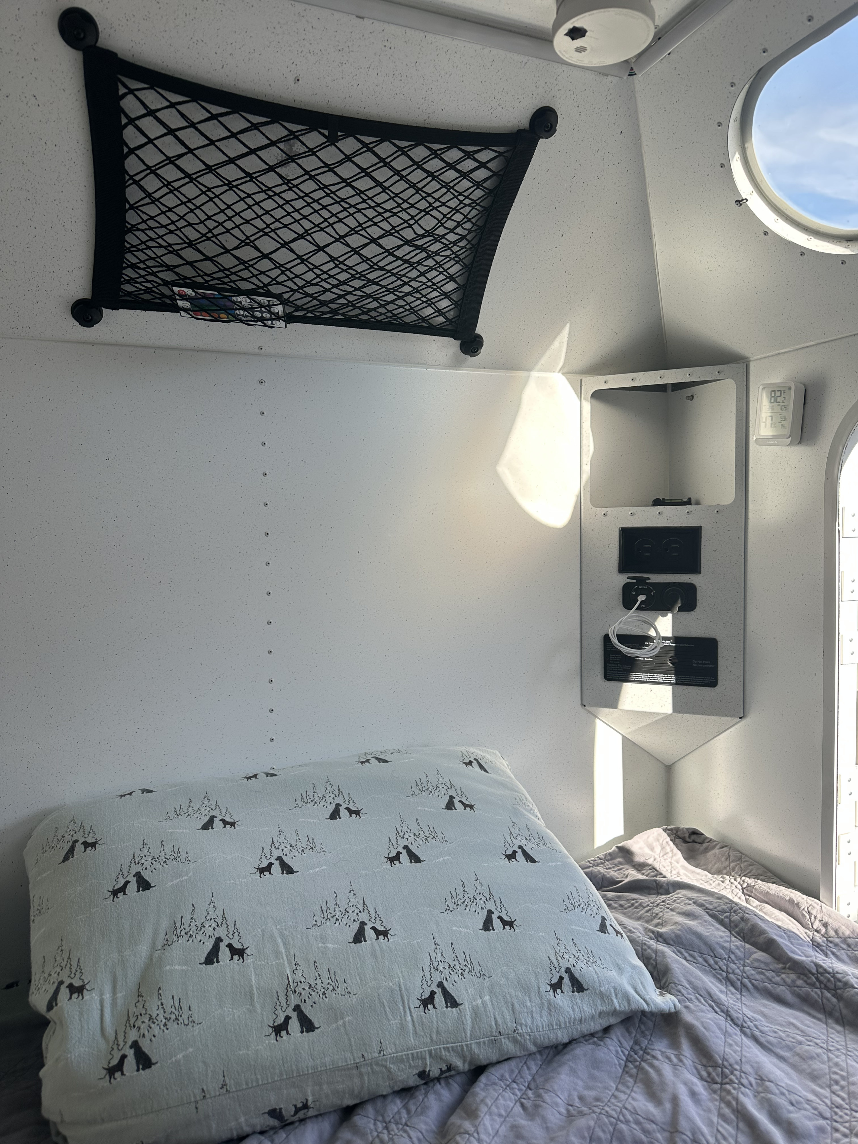 Cozy rv rental interior: bed with patterned pillow, overhead net, power panel, & round window to blue sky.. Mammoth Overland HV 2022