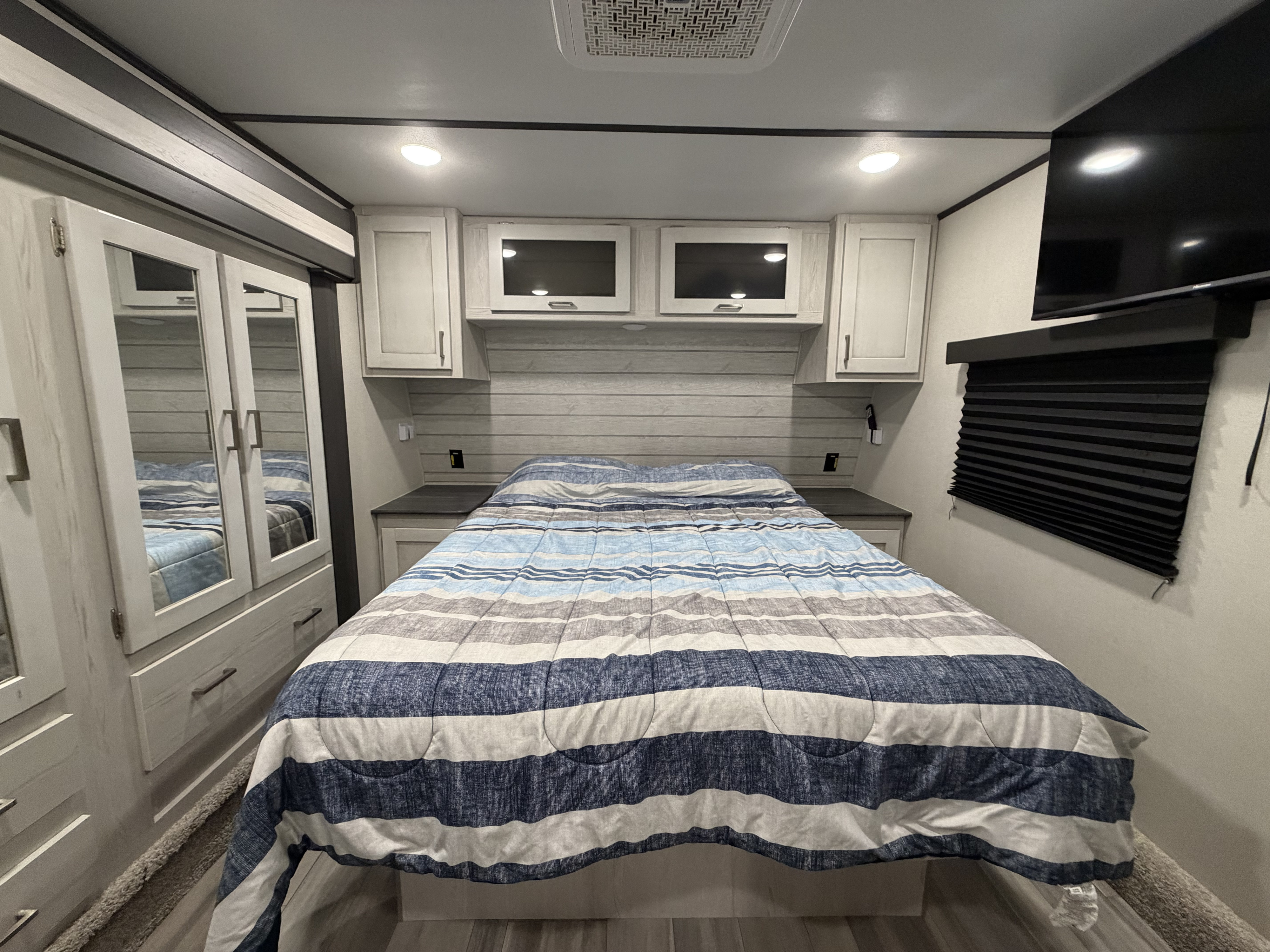 Cozy RV rental bedroom with a striped bed, mirrored wardrobe, overhead cabinets, wall-mounted TV, and pleated window blind.. Keystone RV Sprinter 2022