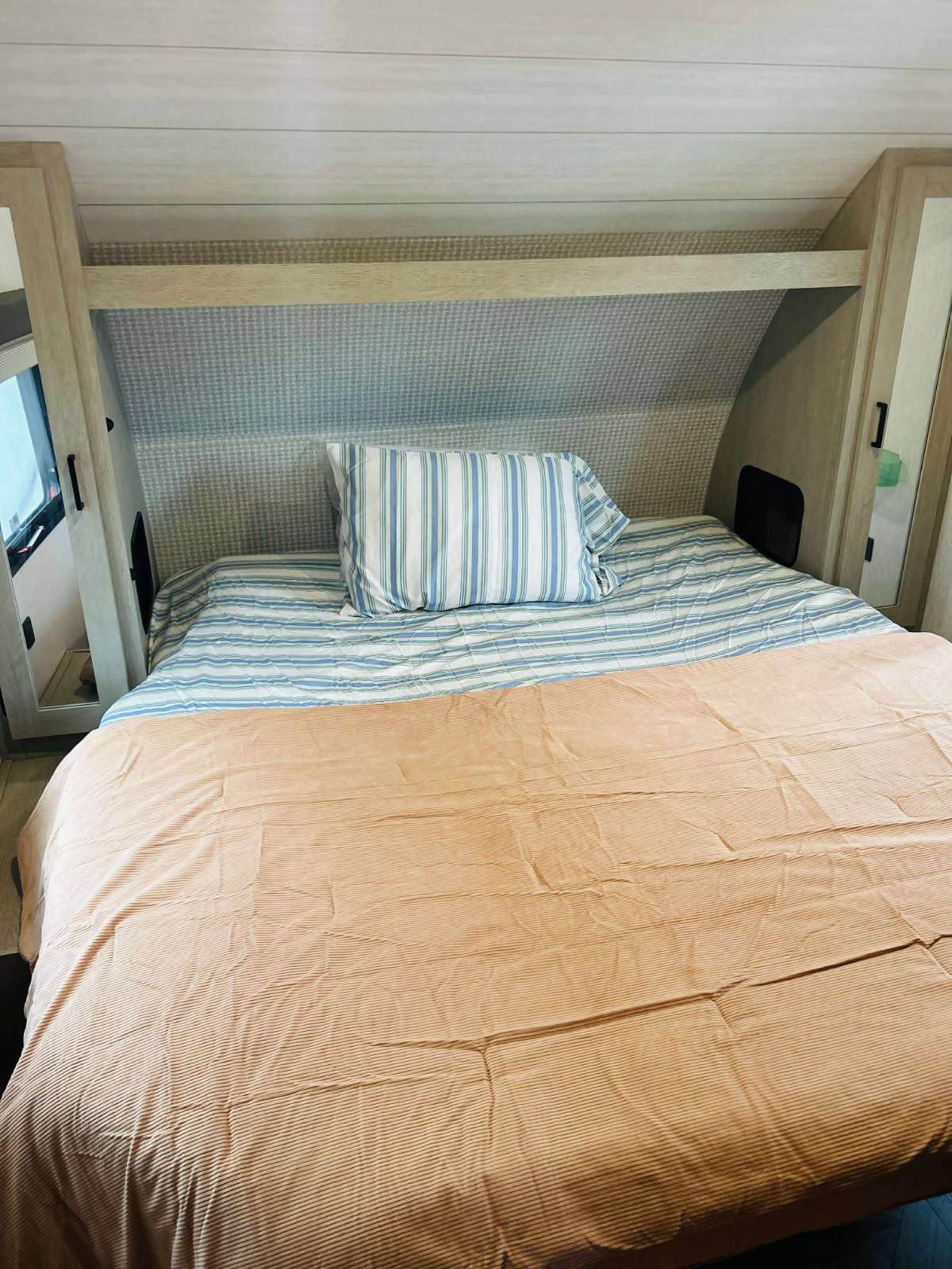 Comfortable RV rental bed with blue and white striped sheets, a peach comforter, and wood-paneled interior.. Forest River Wildwood X-Lite 2024