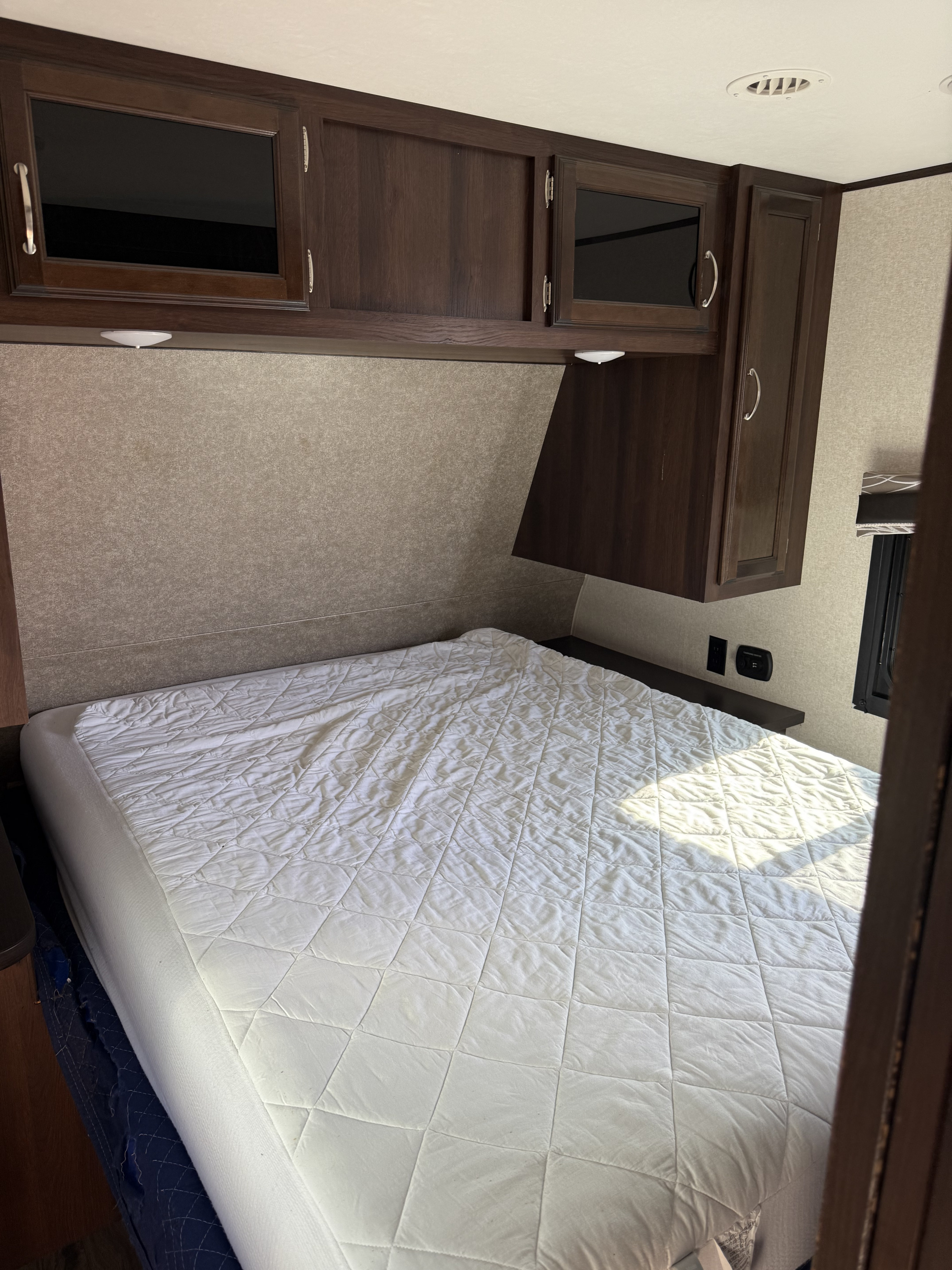 Cozy rv rental bedroom with dark wood overhead cabinets, a quilted mattress, wall outlets, and a window for comfortable travel.. Jayco Jay Flight 2018