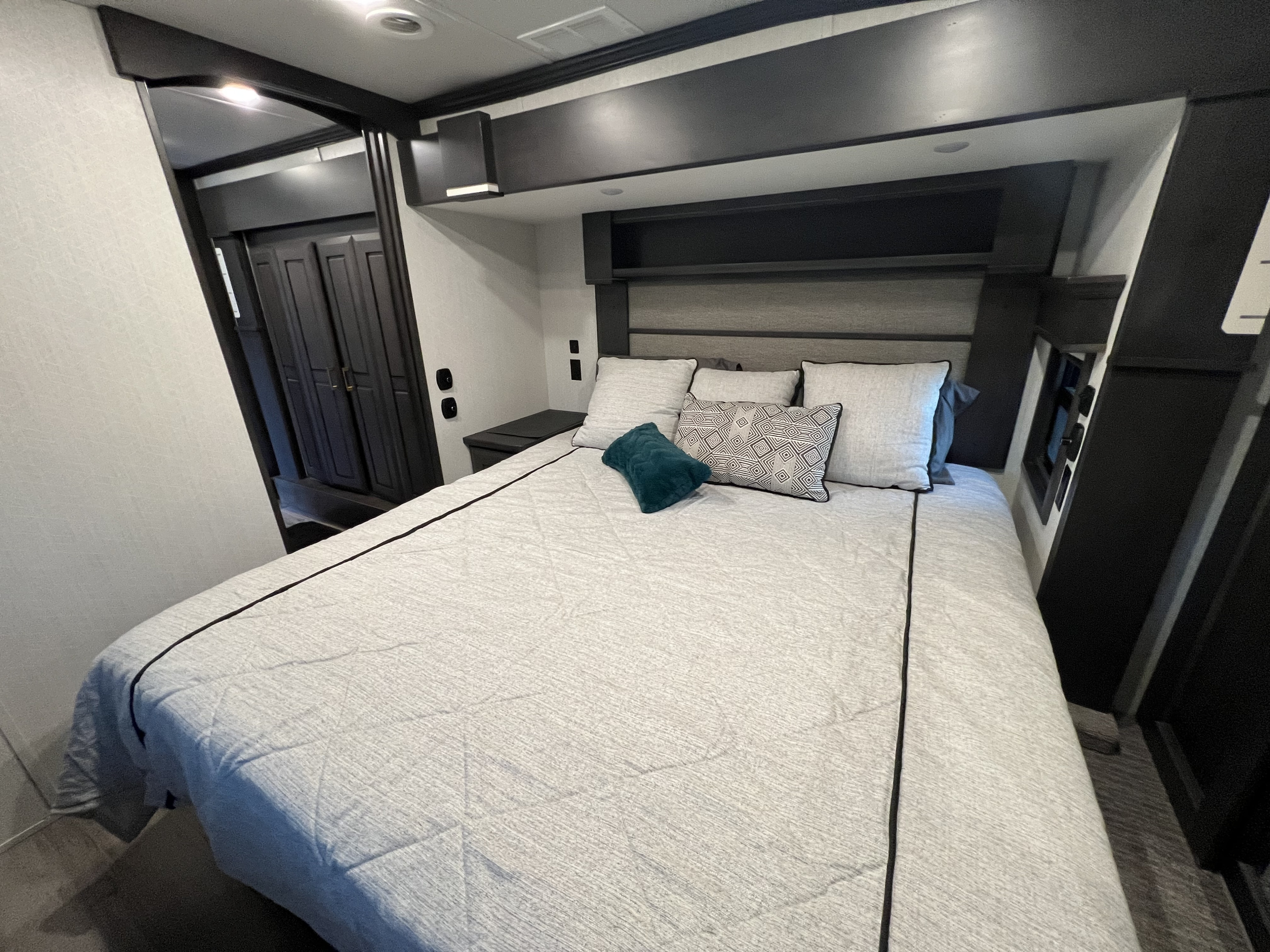 This modern RV rental bedroom features a large gray bed, overhead storage, and built-in nightstands for comfort and convenience.. DRV Mobile Suites 2021