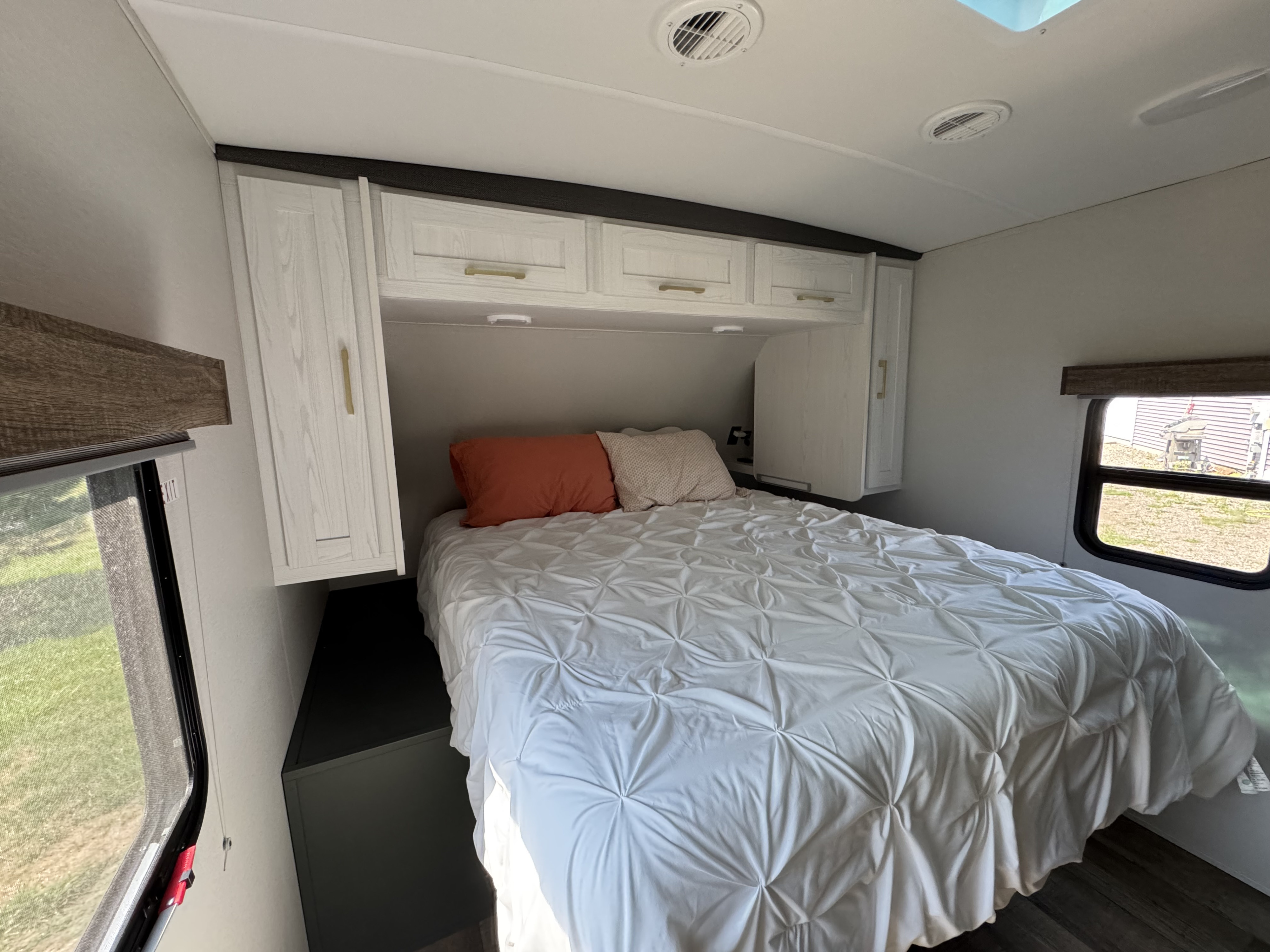 Cozy RV rental bedroom featuring a bed with a white comforter, pillows, overhead cabinets, and windows on both sides.. Heartland RVs 260RB 2024