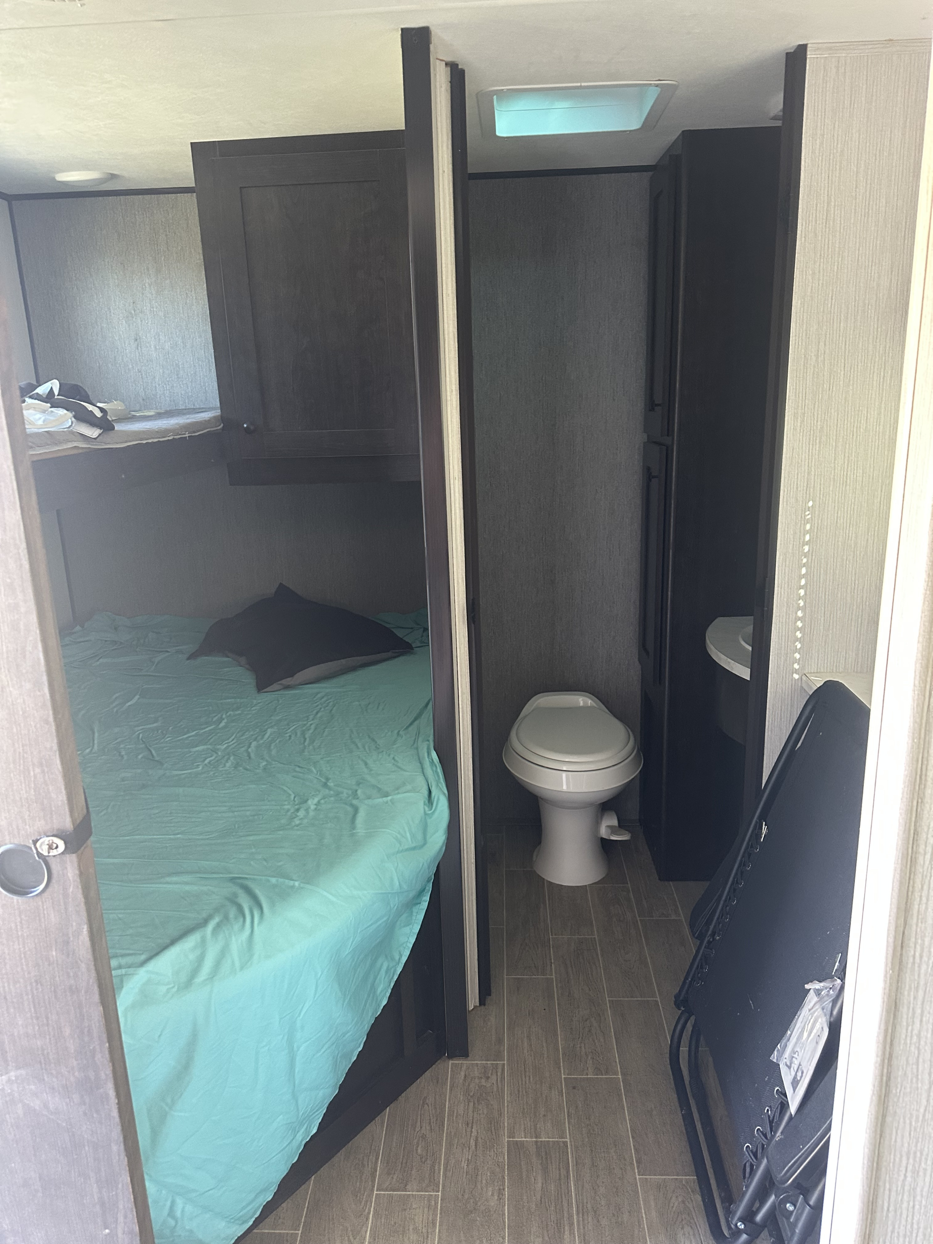 RV rental interior: bunk beds with teal sheets, a toilet, and a folded chair on wood-look floor. Compact living space.. Heartland RVs Trail Runner 2020