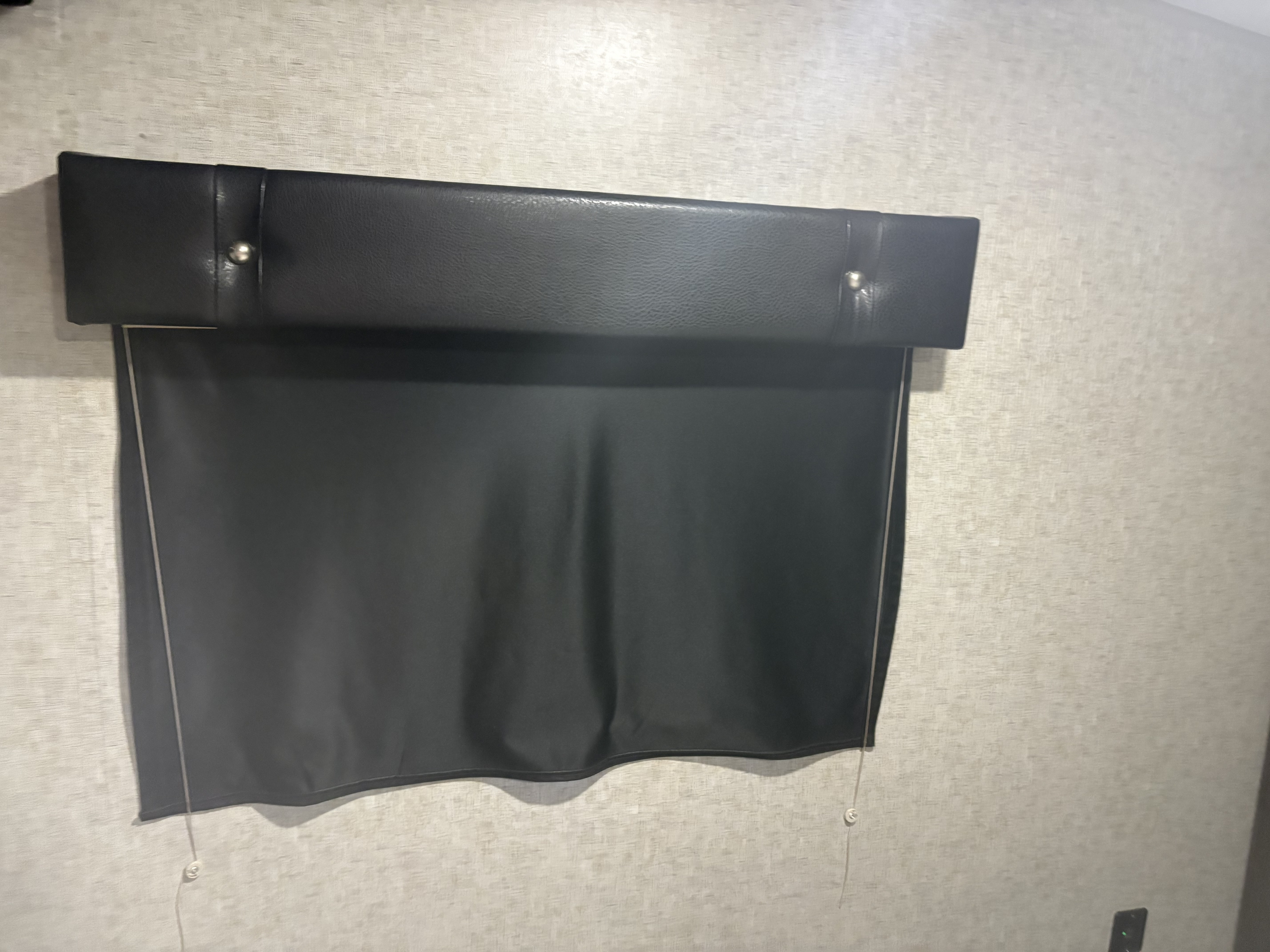 Inside an RV rental: a black pull-down window shade with a faux leather padded valance on a textured wall.. Forest River Cherokee Wolf Pack 2018