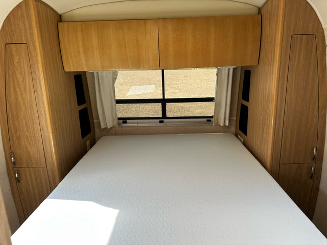 Compact RV rental bedroom with light wood cupboards, a white bed, and a window, offering a cozy and functional travel space.. Airstream Safari 2006