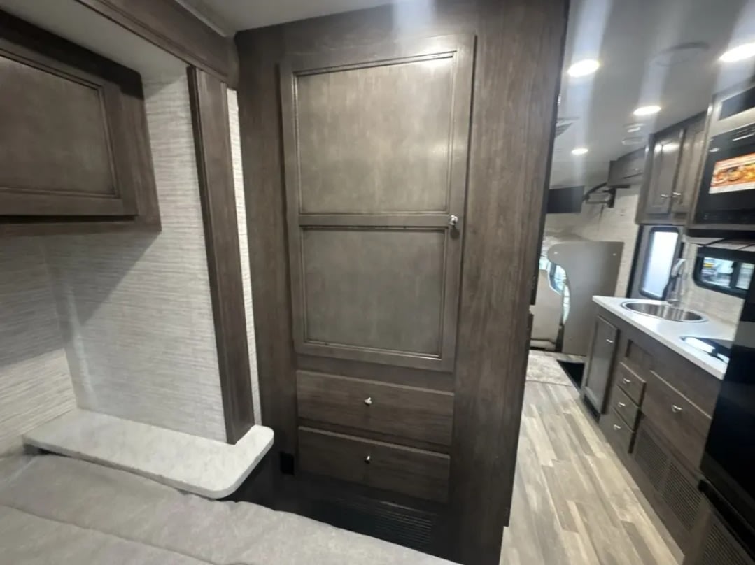 Inside an RV rental: a cozy bedroom wall with dark wood cabinets & drawers, plus a glimpse into the kitchen area with sink & microwave.. Jayco Redhawk SE 2025