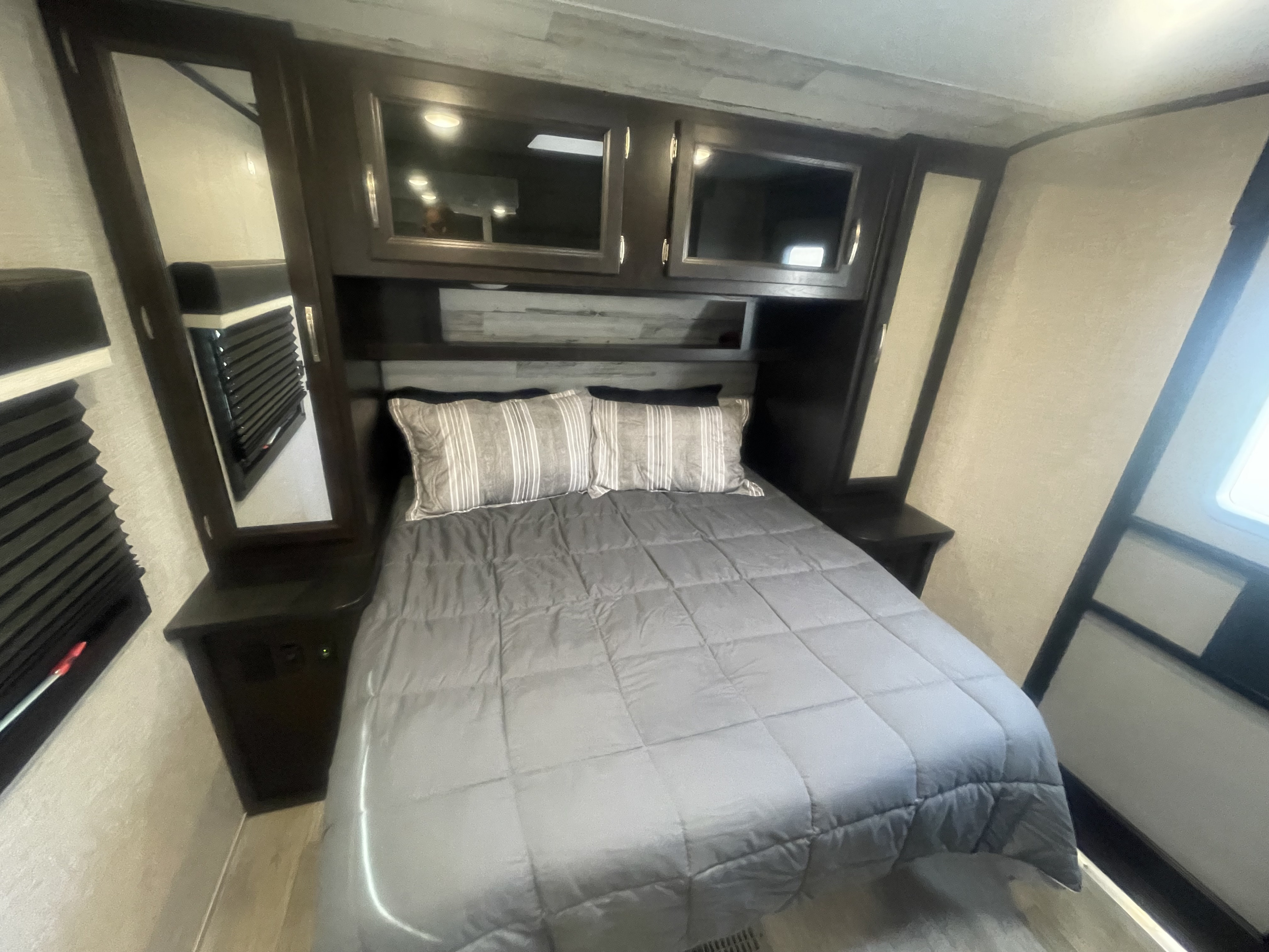 Cozy RV rental bedroom featuring a comfy bed with gray comforter, overhead cabinets, and mirrored details.. Jayco Jay Feather 2022