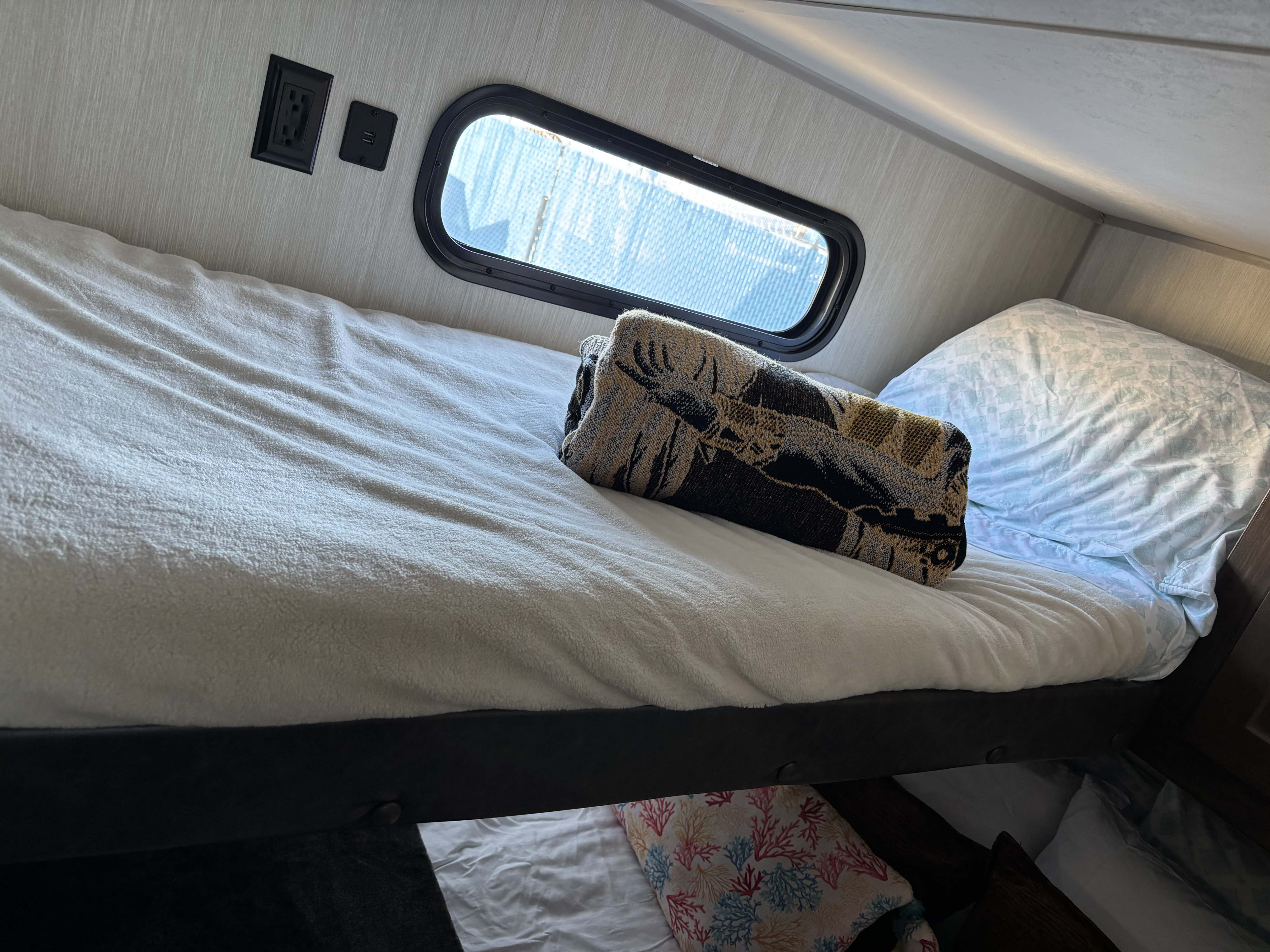 Inside an RV rental, a cozy bunk bed features a window, charging ports, and warm bedding, ideal for travel accommodation.. Forest River Cherokee 2019