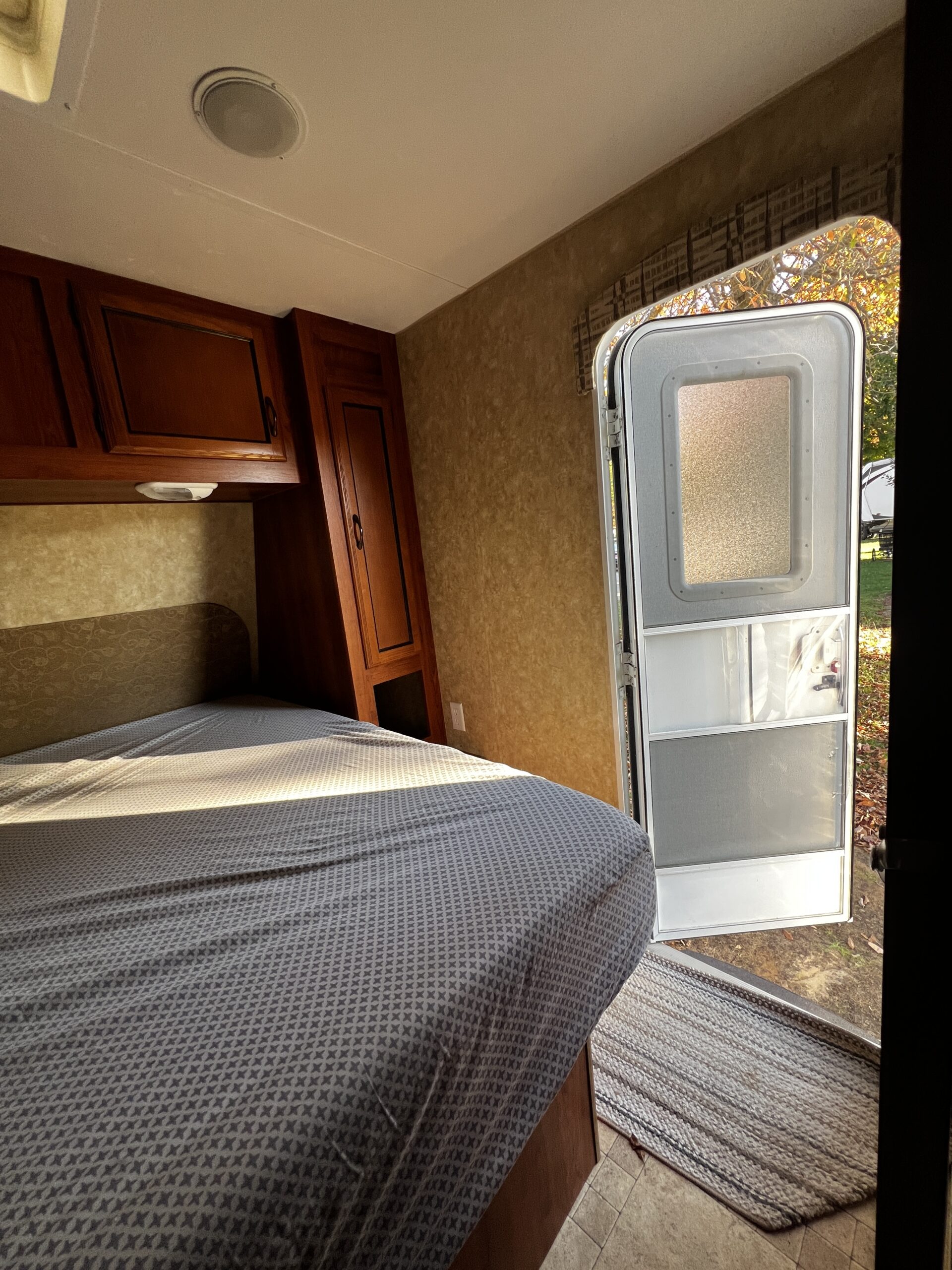 Inside an RV rental, a cozy bedroom features a patterned bed, wood cabinets, and an open door revealing nature outside.. Coachman Catalina 2012