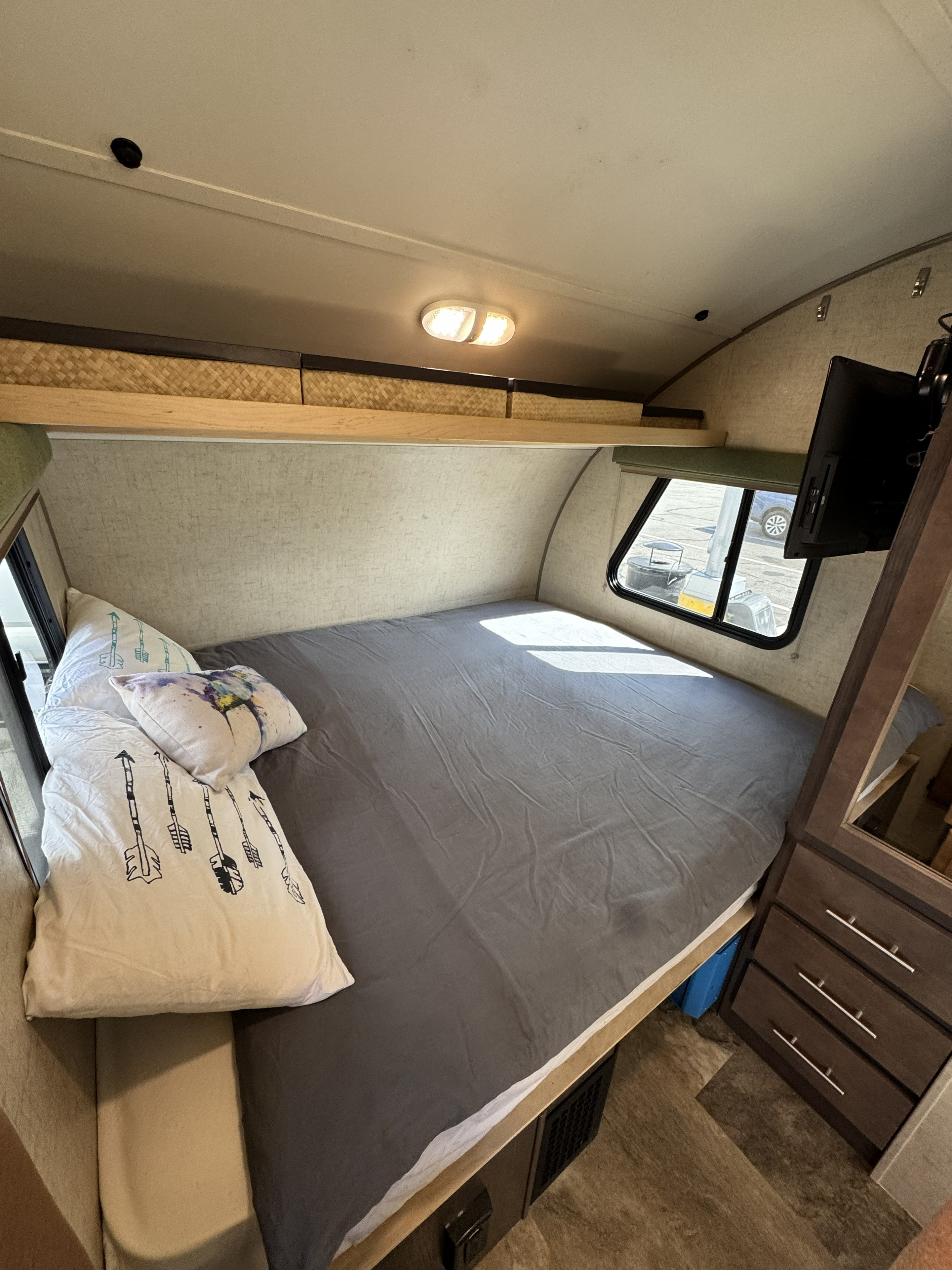 Interior view of an RV rental bedroom with a full bed, gray sheets, patterned pillows, overhead storage, wall-mounted TV, and window.. R-Pod Hood River Edition 2015