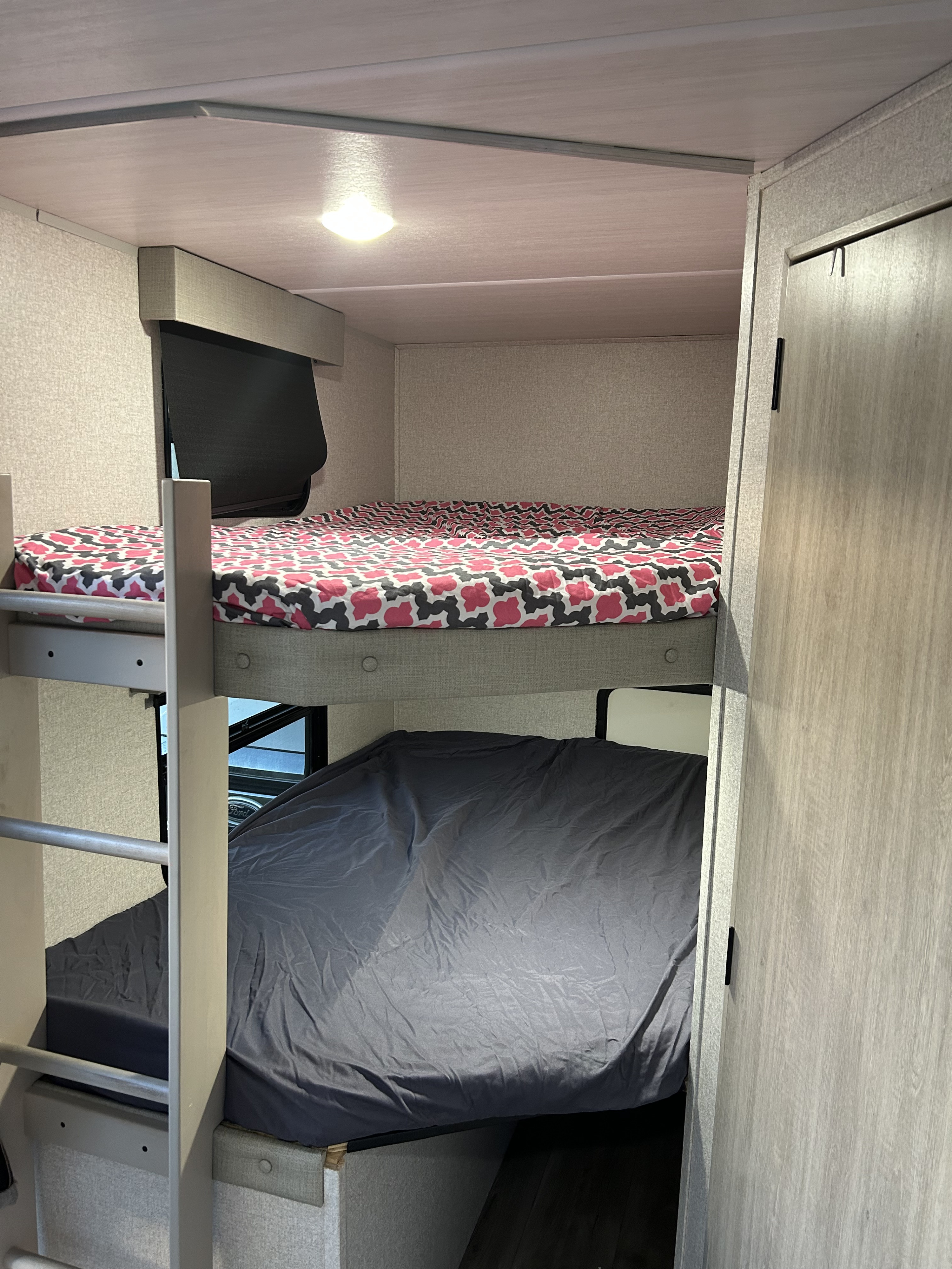 An rv rental's compact interior featuring bunk beds. The top bed has a pink/grey patterned sheet, the bottom a dark grey.. East to West Alta 2023