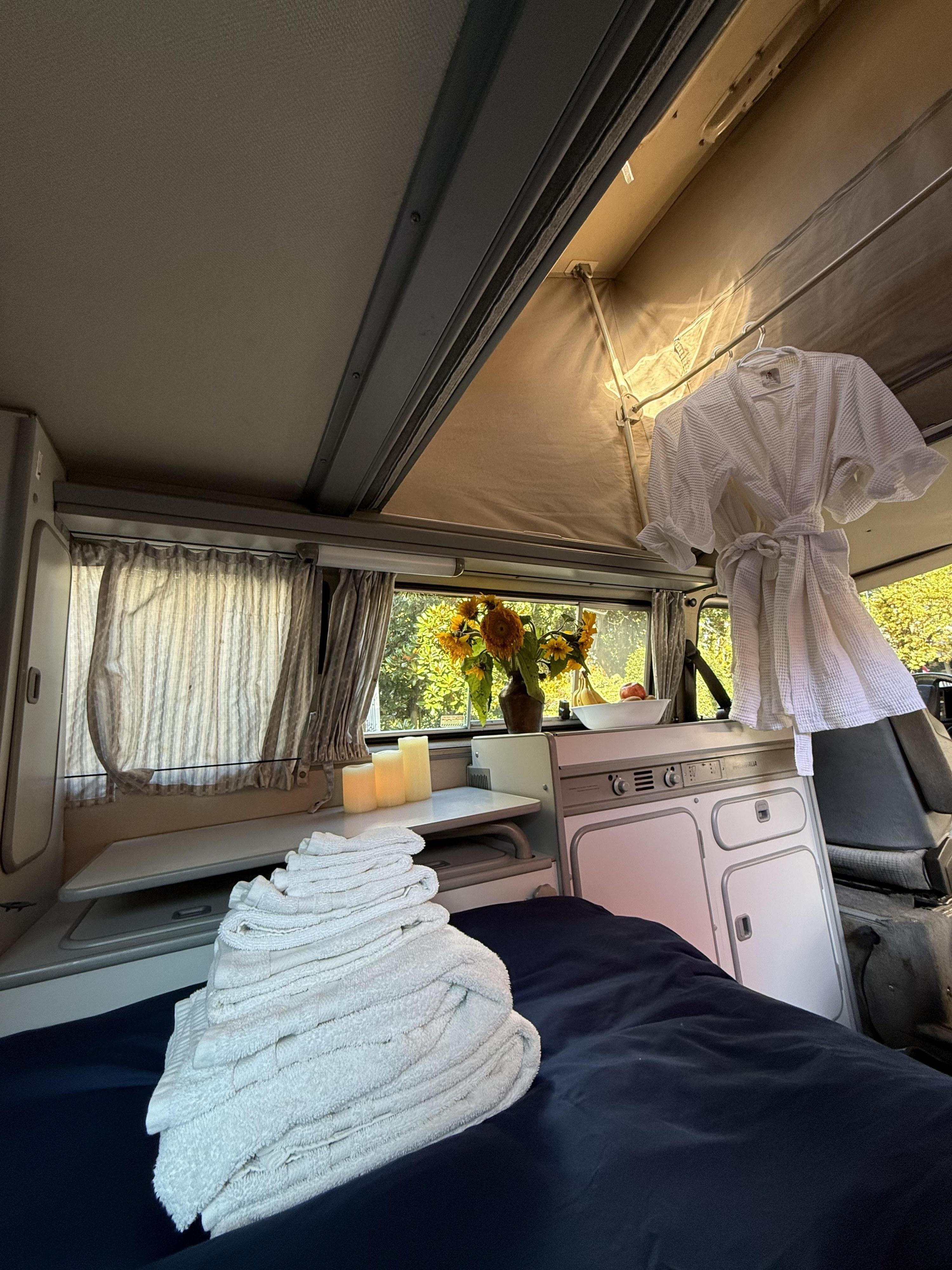 Cozy rv rental interior featuring a bed with fresh towels, sunflowers, and a kitchenette, perfect for a comfortable road trip.. Volkswagen T3 Westfalia 1991