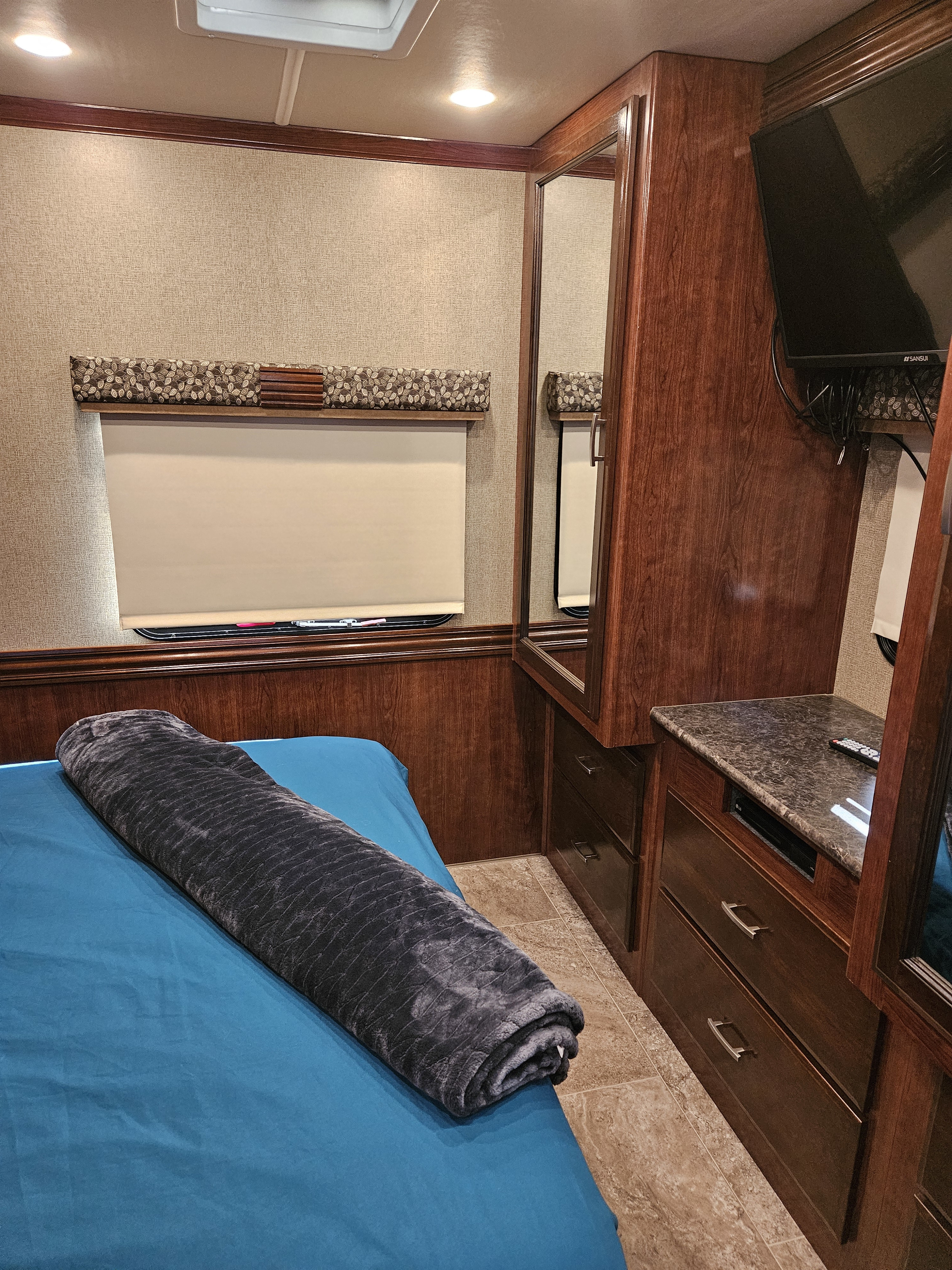 Cozy RV rental bedroom featuring a bed with blue sheets & grey blanket, window, wood cabinetry, TV & mirror.. Holiday Rambler Vesta 2018
