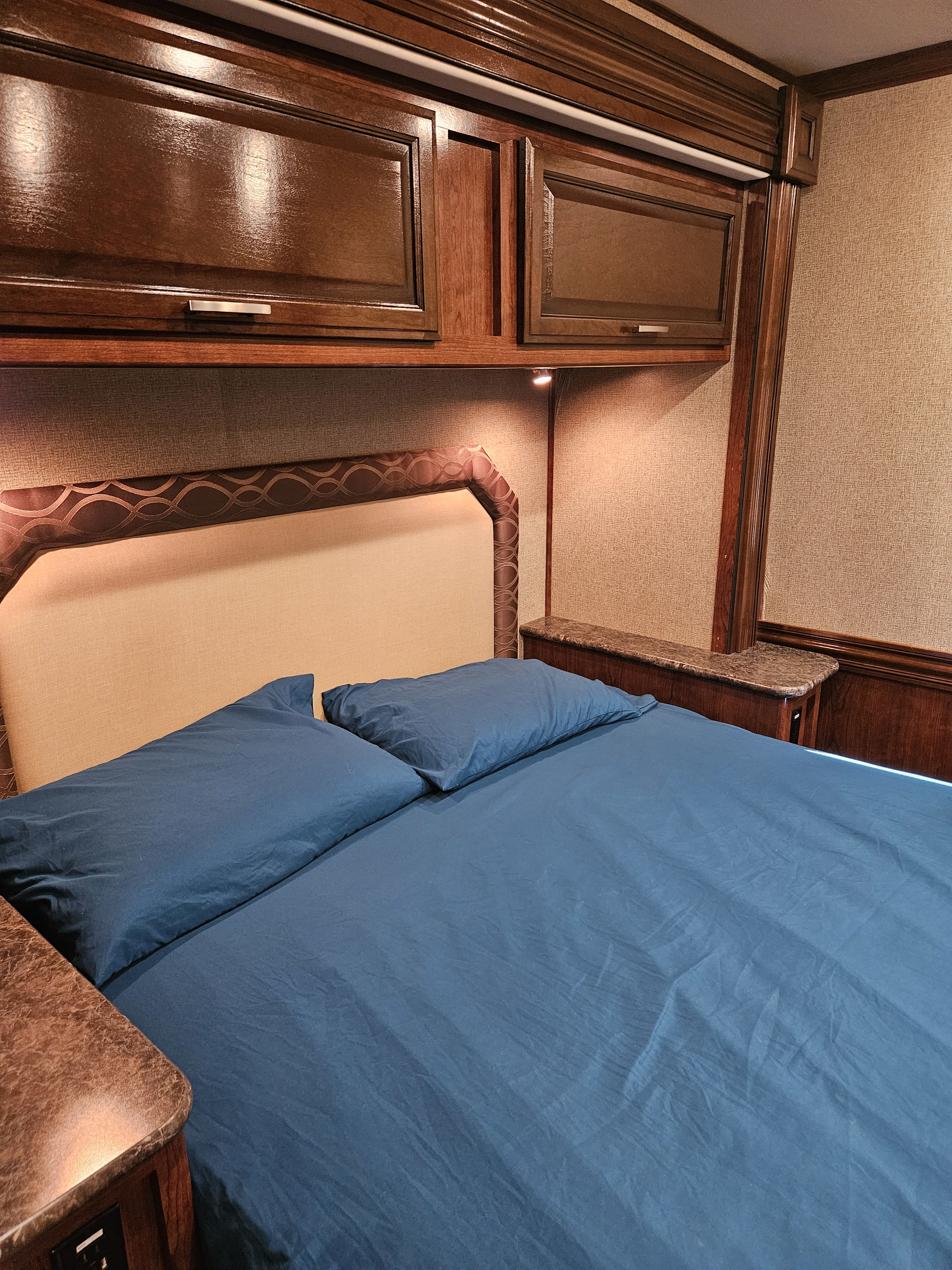 Cozy RV rental bedroom featuring a bed with blue bedding, overhead wooden cabinets, and task lighting.. Holiday Rambler Vesta 2018