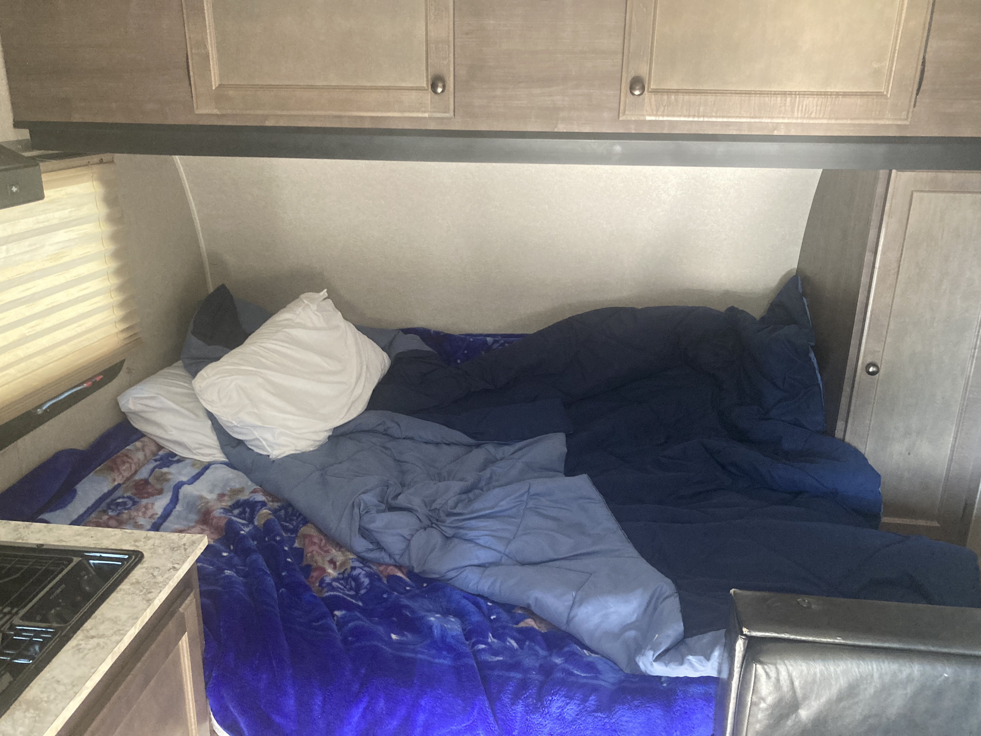 Unmade bed in a compact RV rental sleeping area, with cabinets, a window, and a kitchen counter visible.. Sunset Park RV Sun Lite 2021
