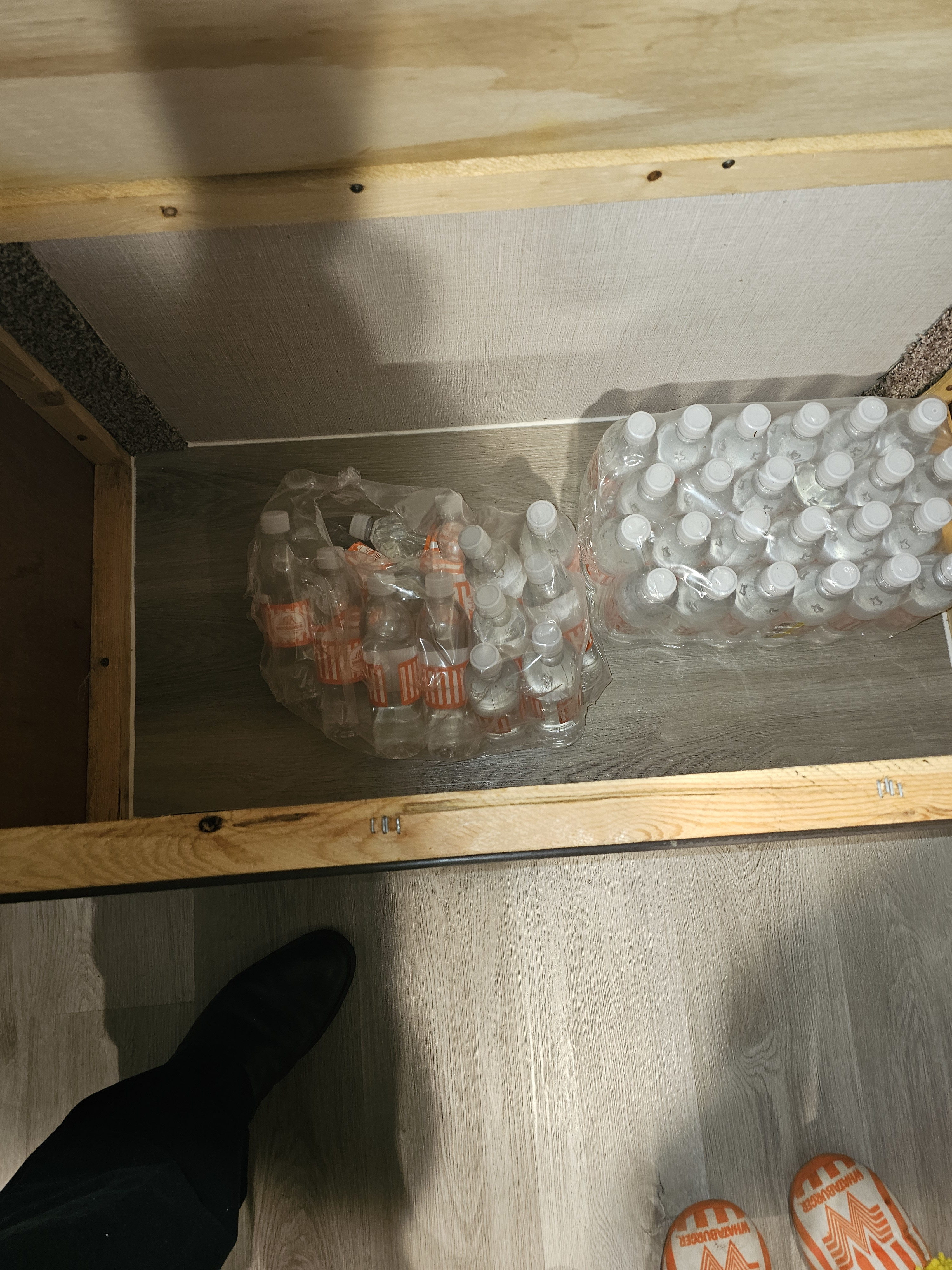 RV rental storage: cases of bottled drinks, including Whataburger, under a bed frame. Feet in slipper & shoe on laminate.. Forest River Palomino Palomini 2018