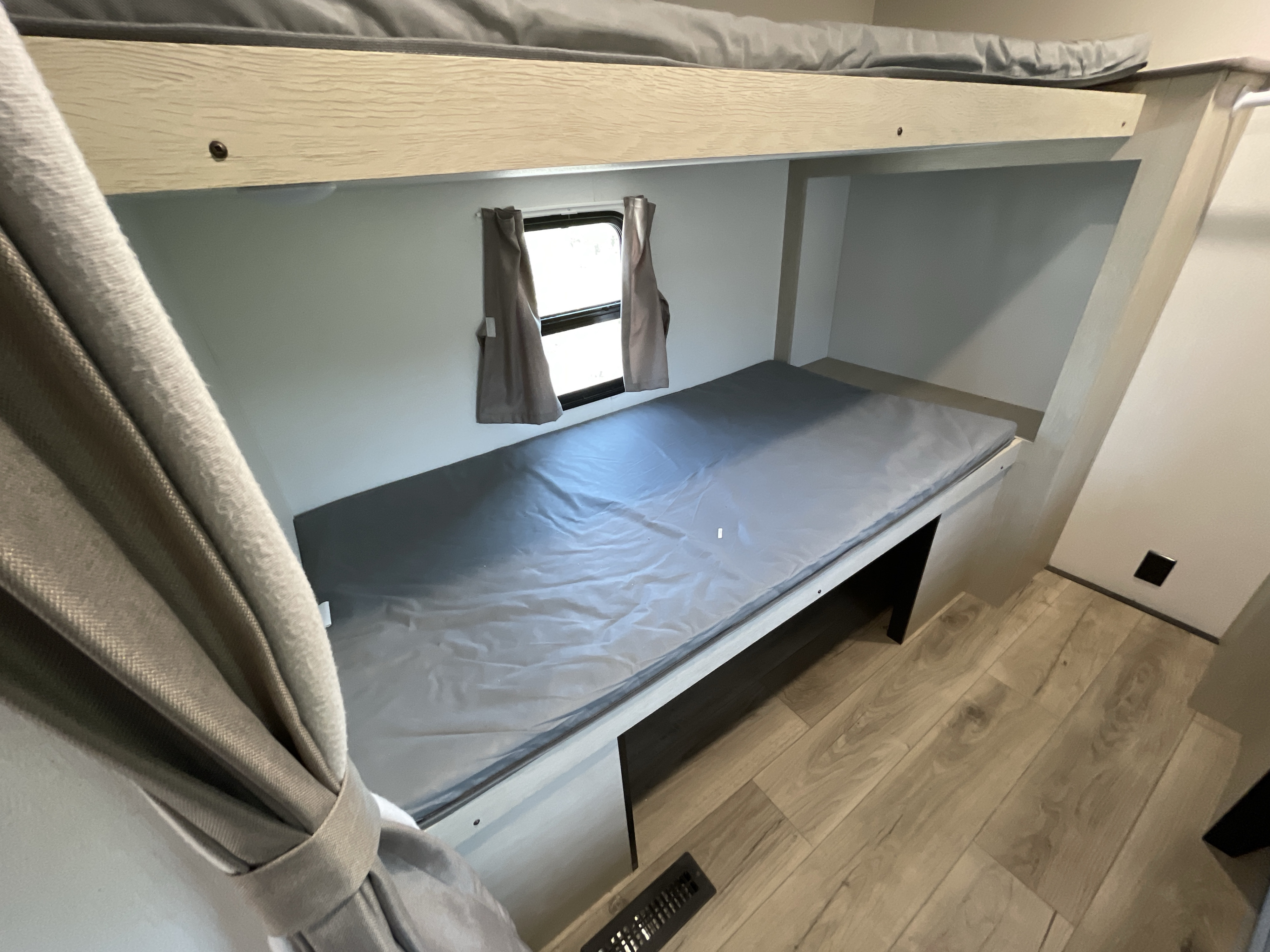 Cozy lower bunk bed in an RV rental, featuring a grey mattress, small window with curtains, and light wood flooring.. Coachmen Shasta 31QB 2025