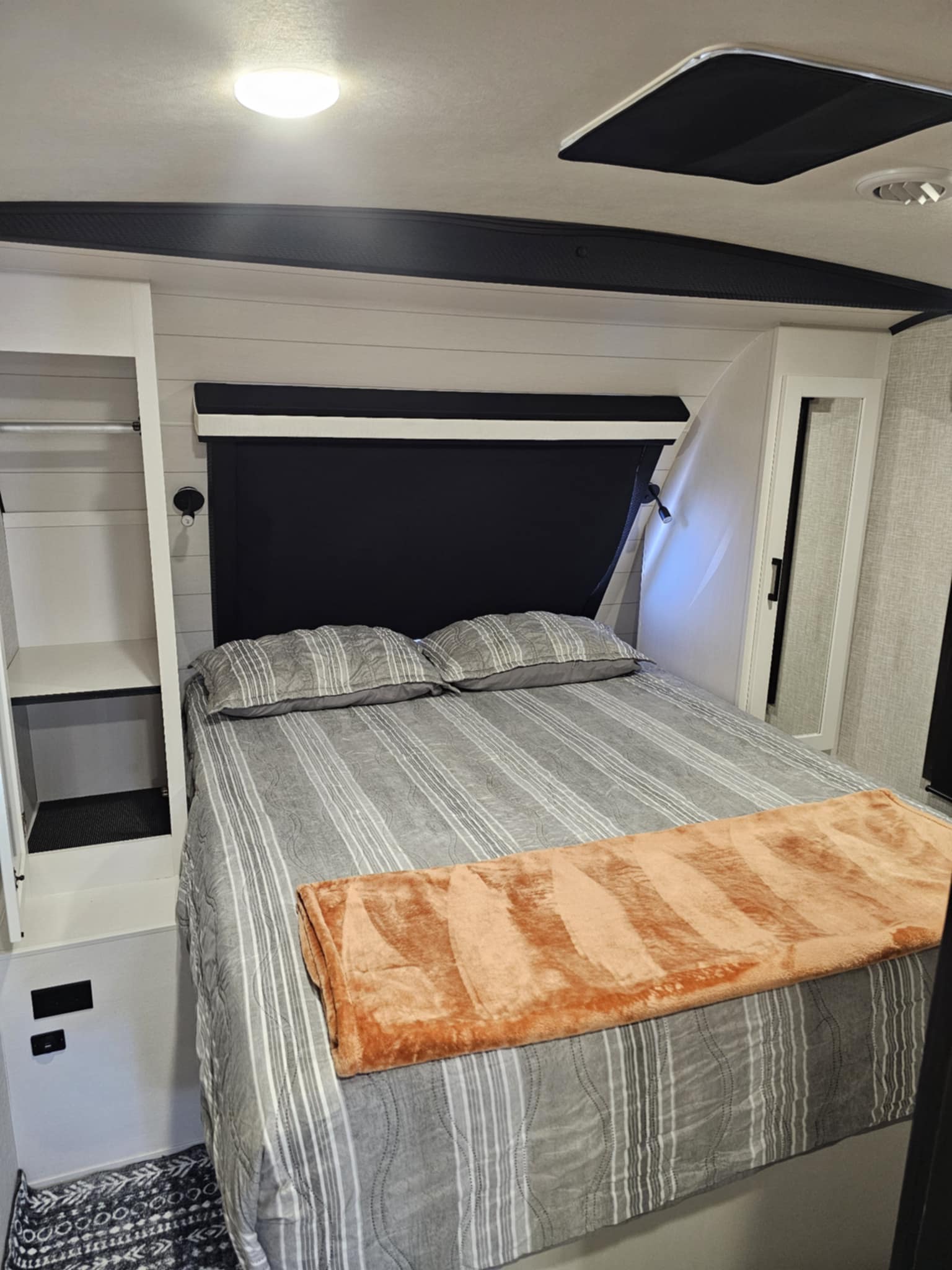 Cozy RV rental bedroom. Features a gray striped bed, orange blanket, built-in storage, and wall sconces for reading.. Jayco White Hawk 2024