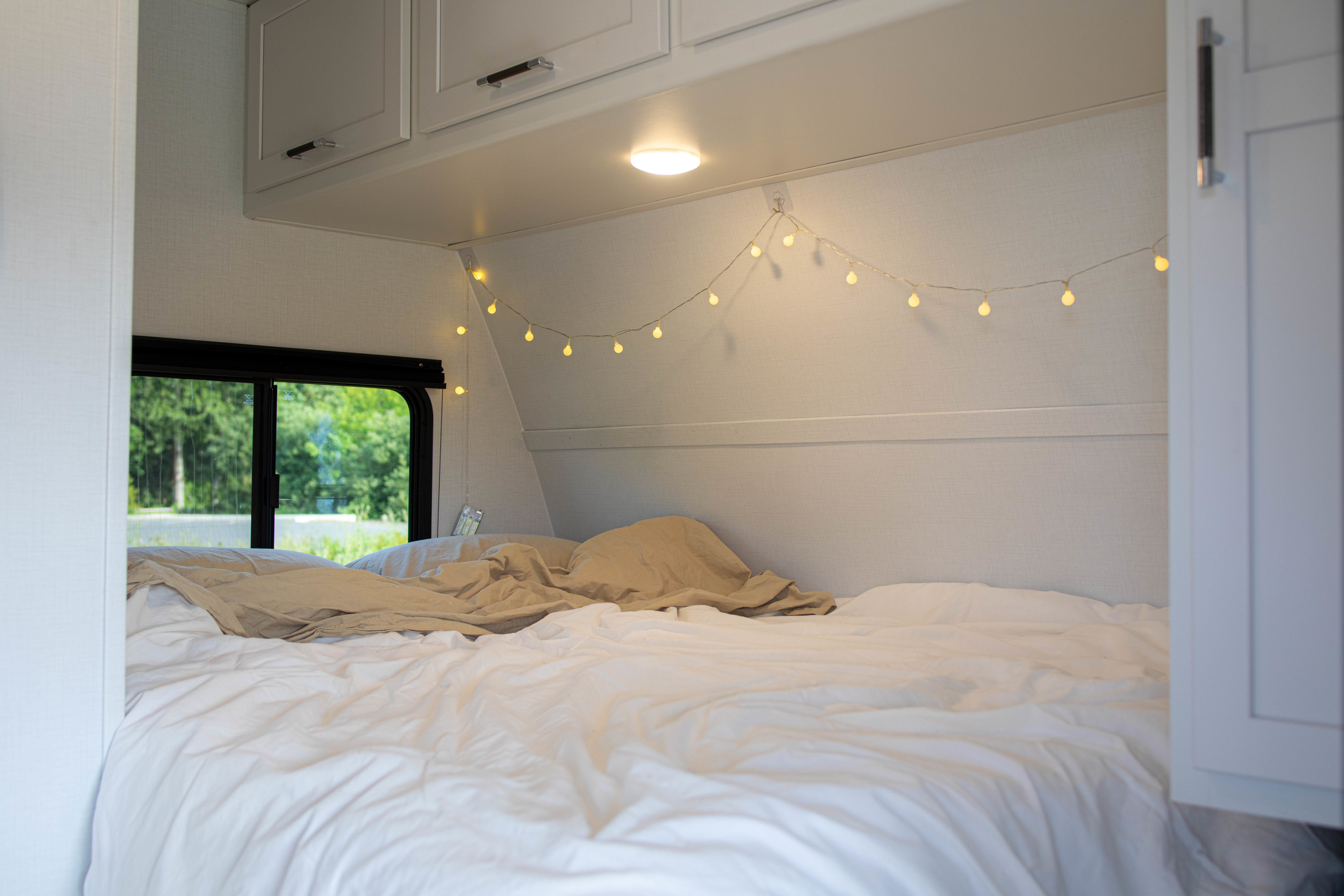 Cozy RV rental bedroom featuring a comfortable bed with string lights, overhead cabinets, and a window showcasing green trees.. Keystone RV Bullet Classic 2025