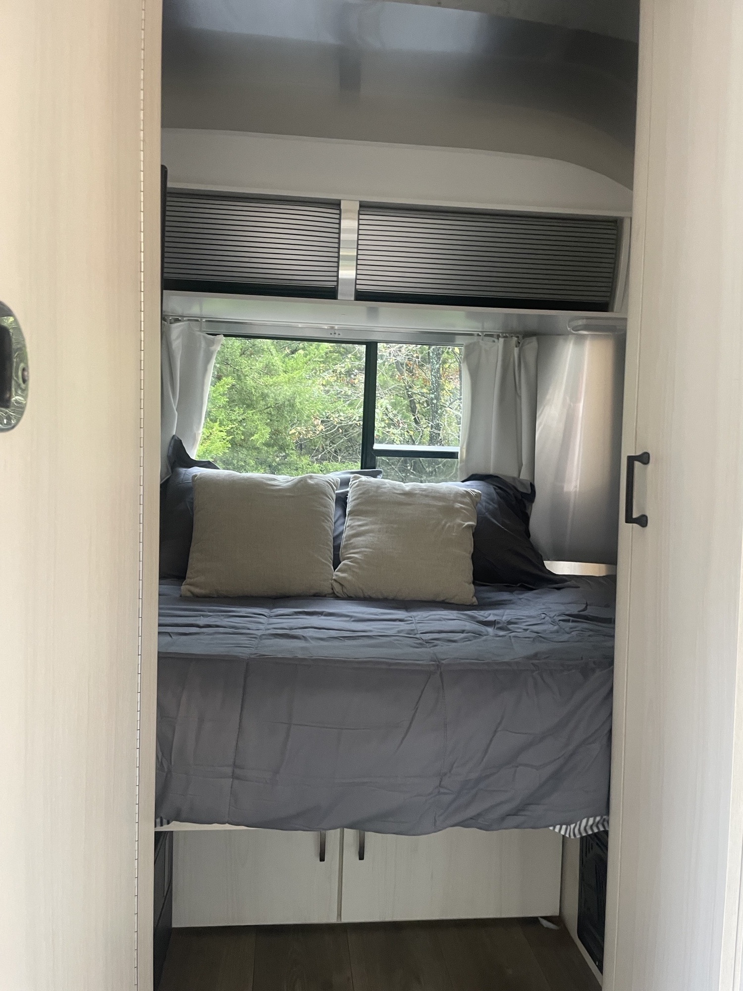 Relaxing rv rental bedroom interior, featuring a comfy bed with grey bedding, pillows, and a window view of lush trees.. Airstream Sport 2019