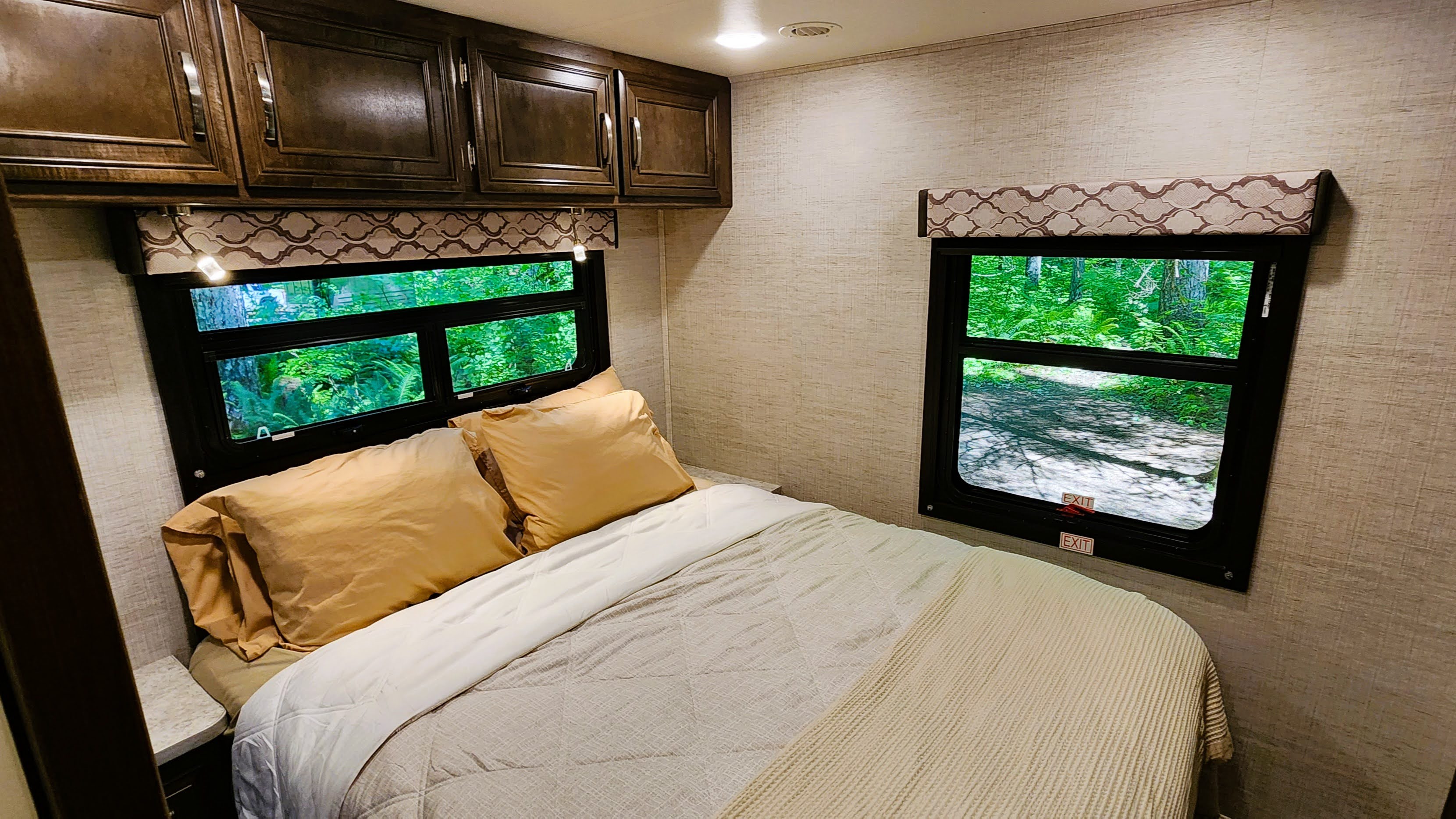 RV rental bedroom: cozy bed with golden pillows, dark cabinets, and scenic windows revealing lush greenery outside.. Jayco 31F 2021