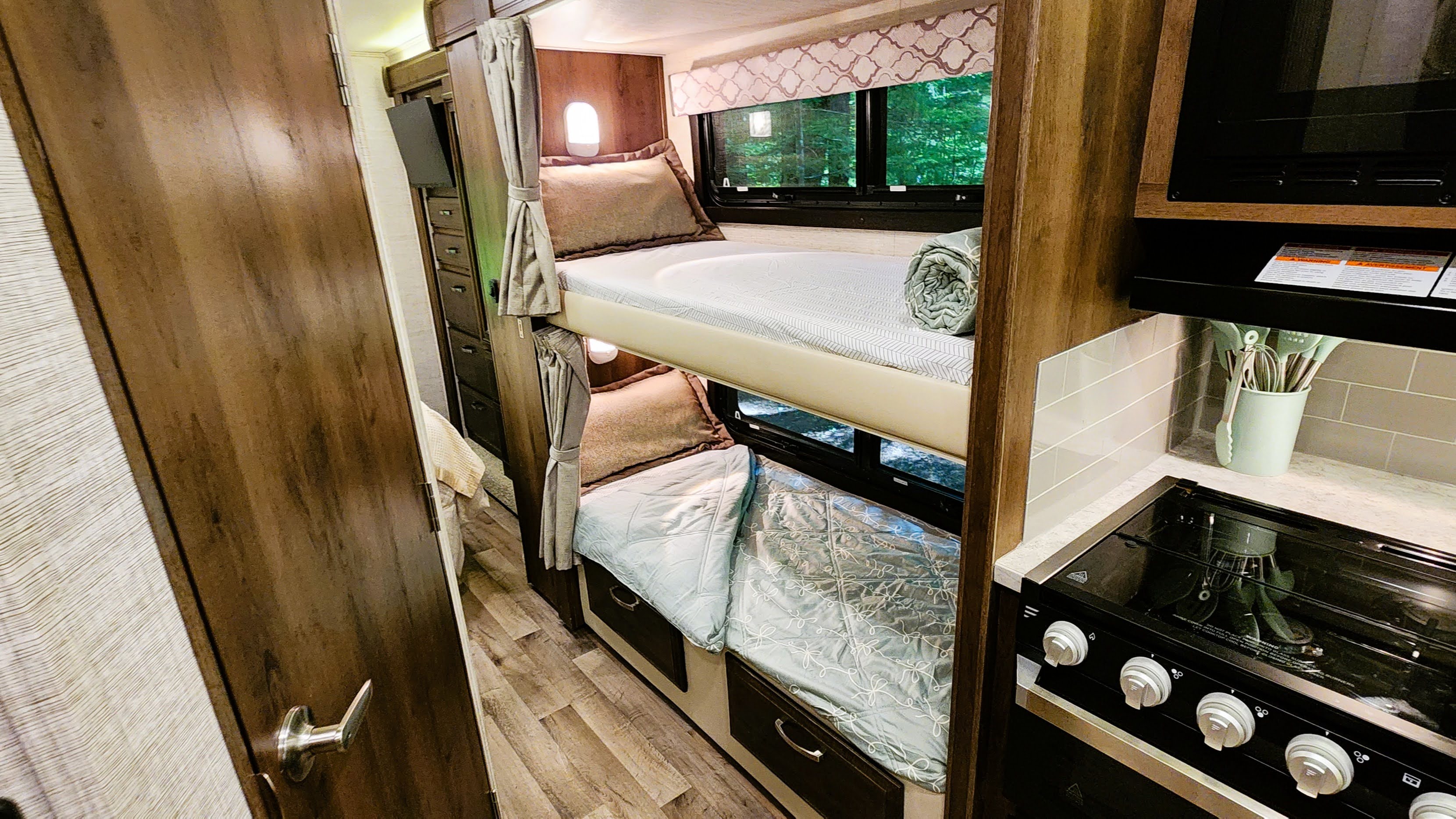 RV rental interior featuring cozy bunk beds next to a compact kitchen with a stove, visible through a wooden door. Ideal for travel.. Jayco 31F 2021