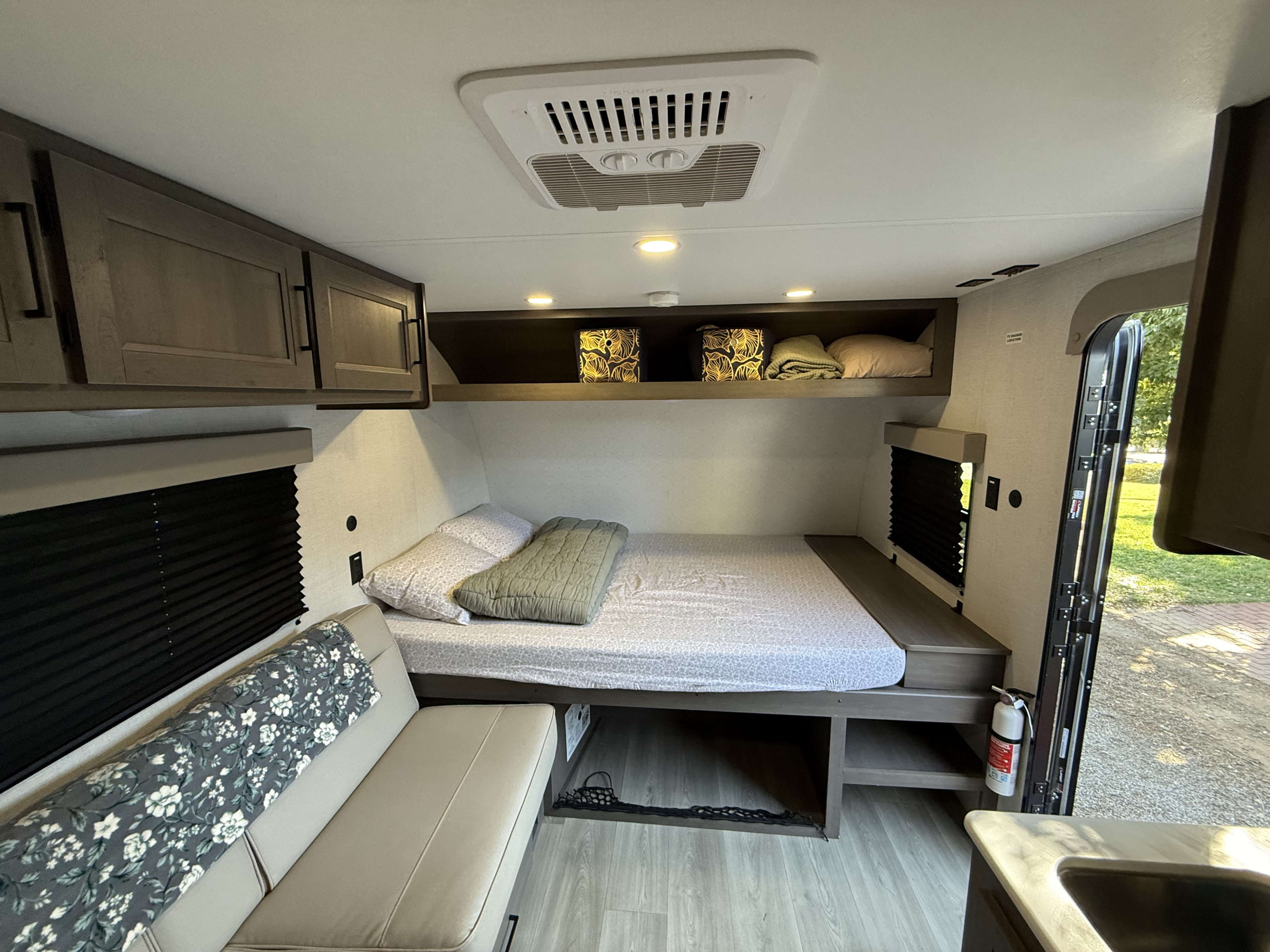 This cozy rv rental interior features a bed on the right, a couch on the left, overhead cabinets, and an AC unit on the ceiling.. Grand Design Transcend One 2025