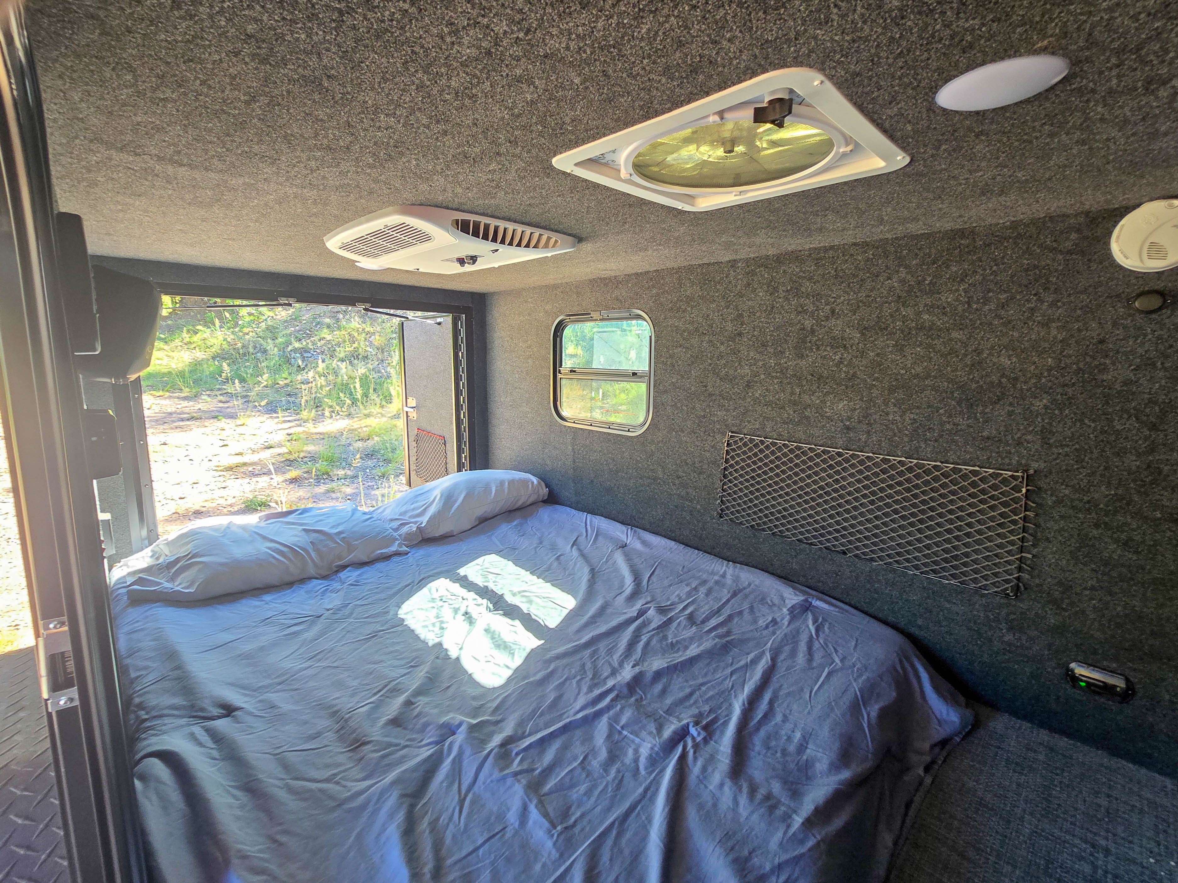 An RV rental's cozy bedroom features a bed, window, ceiling fan, AC unit, and storage net. A door opens to nature.. InTech RV Flyer 2024