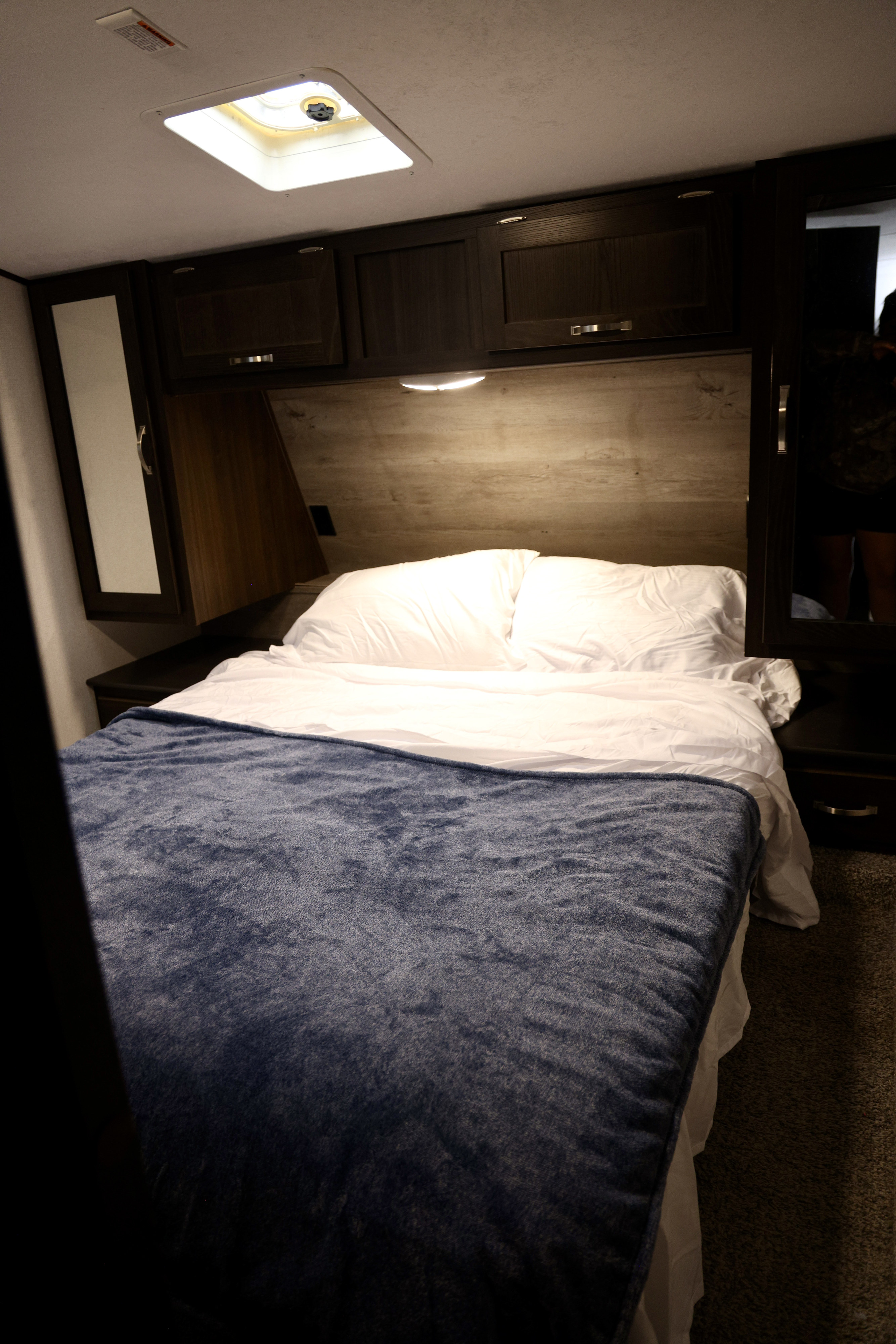 Interior of a cozy RV rental bedroom showing a bed with a blue blanket, white pillows, dark overhead cabinets, and a ceiling light.. Keystone RV Springdale 2020