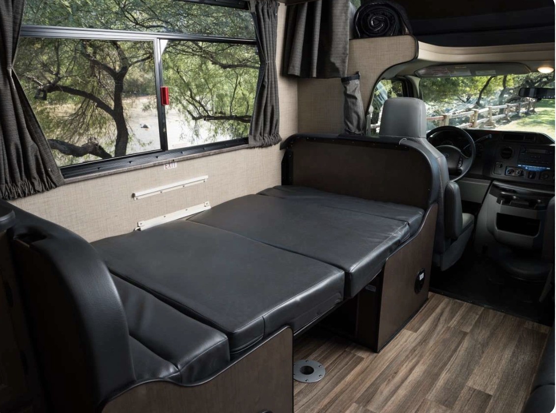 Inside an **rv rental**: a flexible bed/seating area with a large window overlooking nature, leading to the cockpit.. Thor Majestic 2019