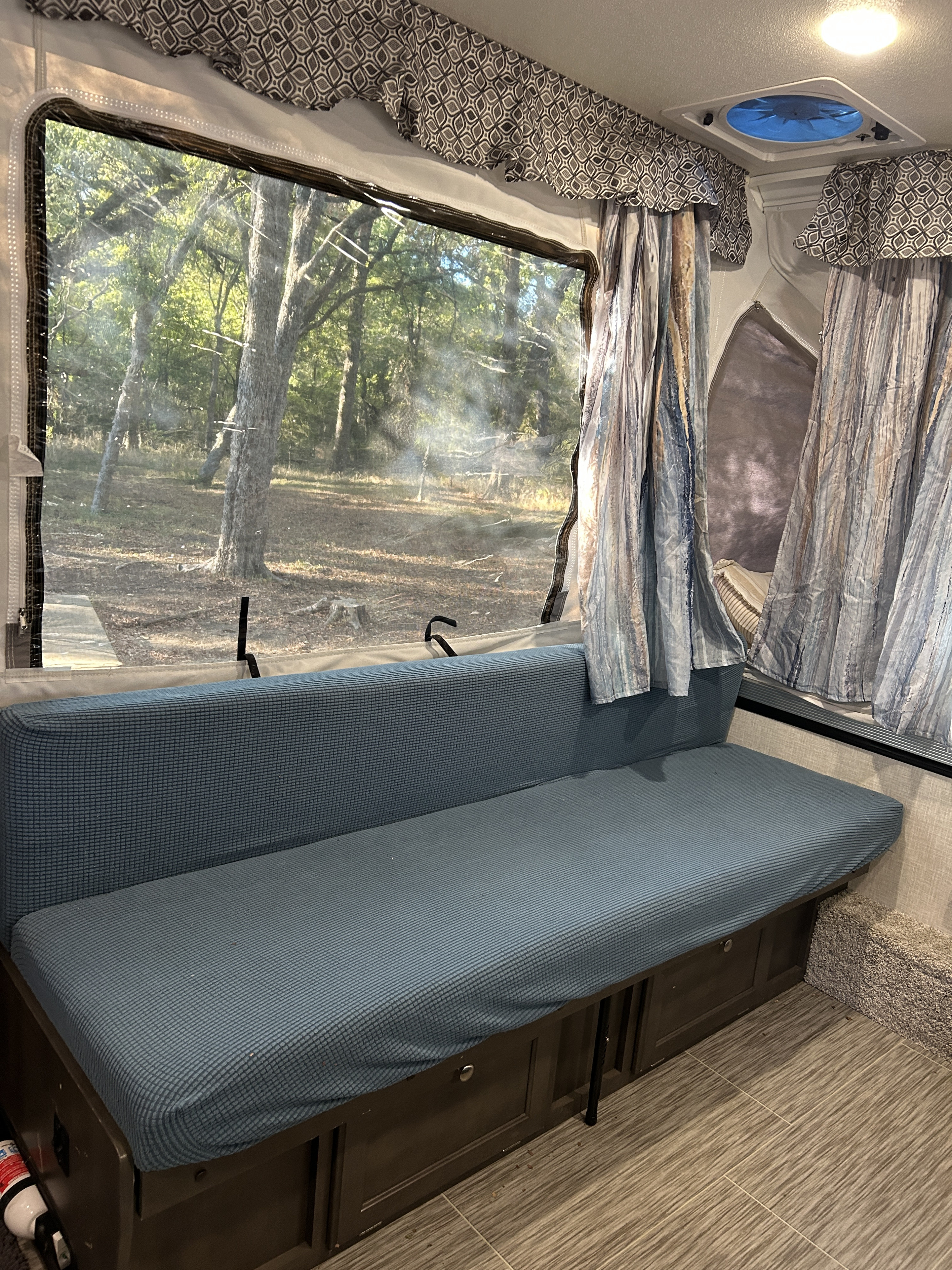 RV rental interior featuring a blue bench, forest view through clear window, patterned valances, and a ceiling fan.. Forest River Flagstaff M.A.C. 2021