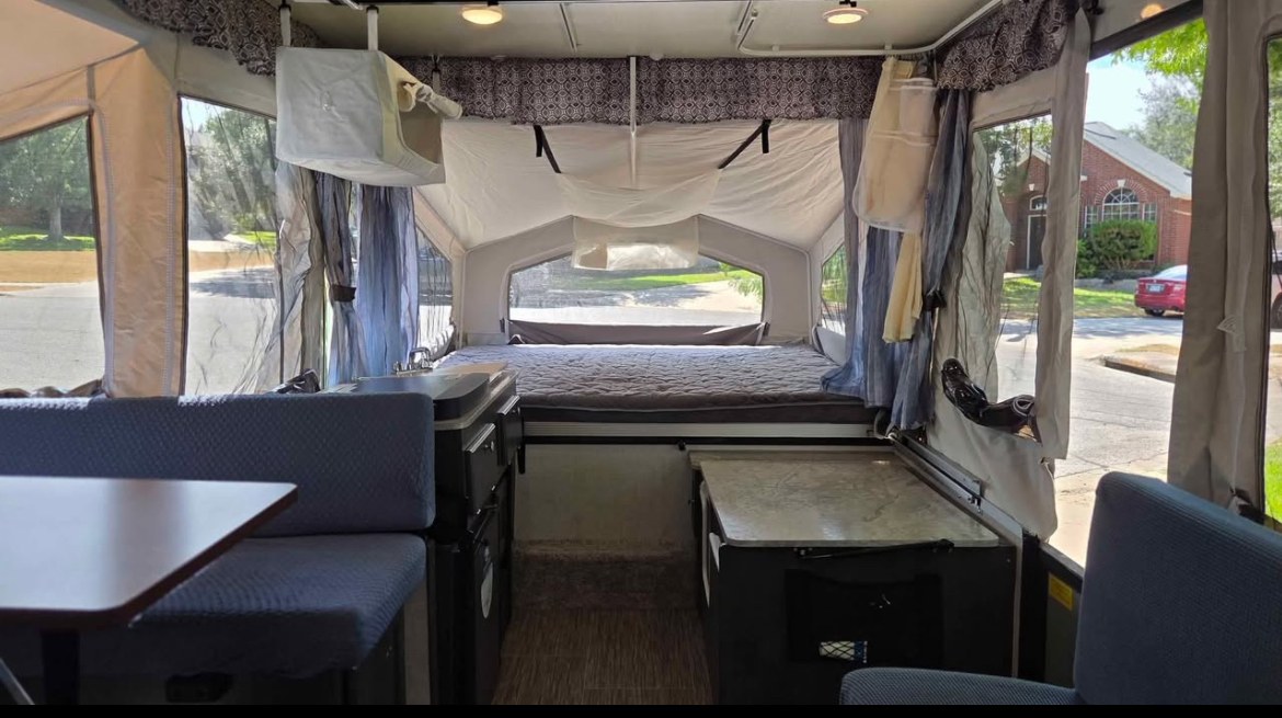 Inside a compact rv rental pop-up camper, featuring a cozy dinette, kitchen, and a bed, with windows showing a street view.. Forest River Flagstaff M.A.C. 2021