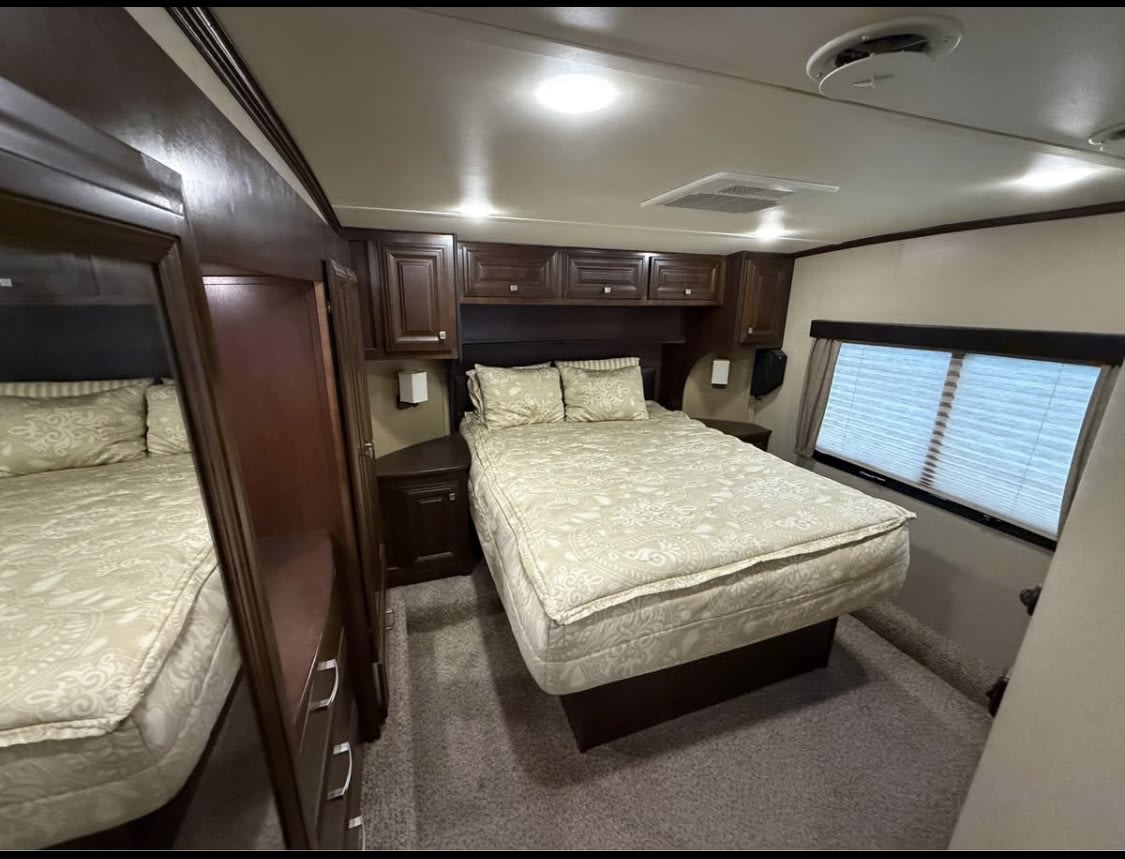 Comfortable RV rental bedroom featuring two beds, dark wood cabinets, bright lighting, and a large window with blinds.. Dutchmen Voltage V3200 2014