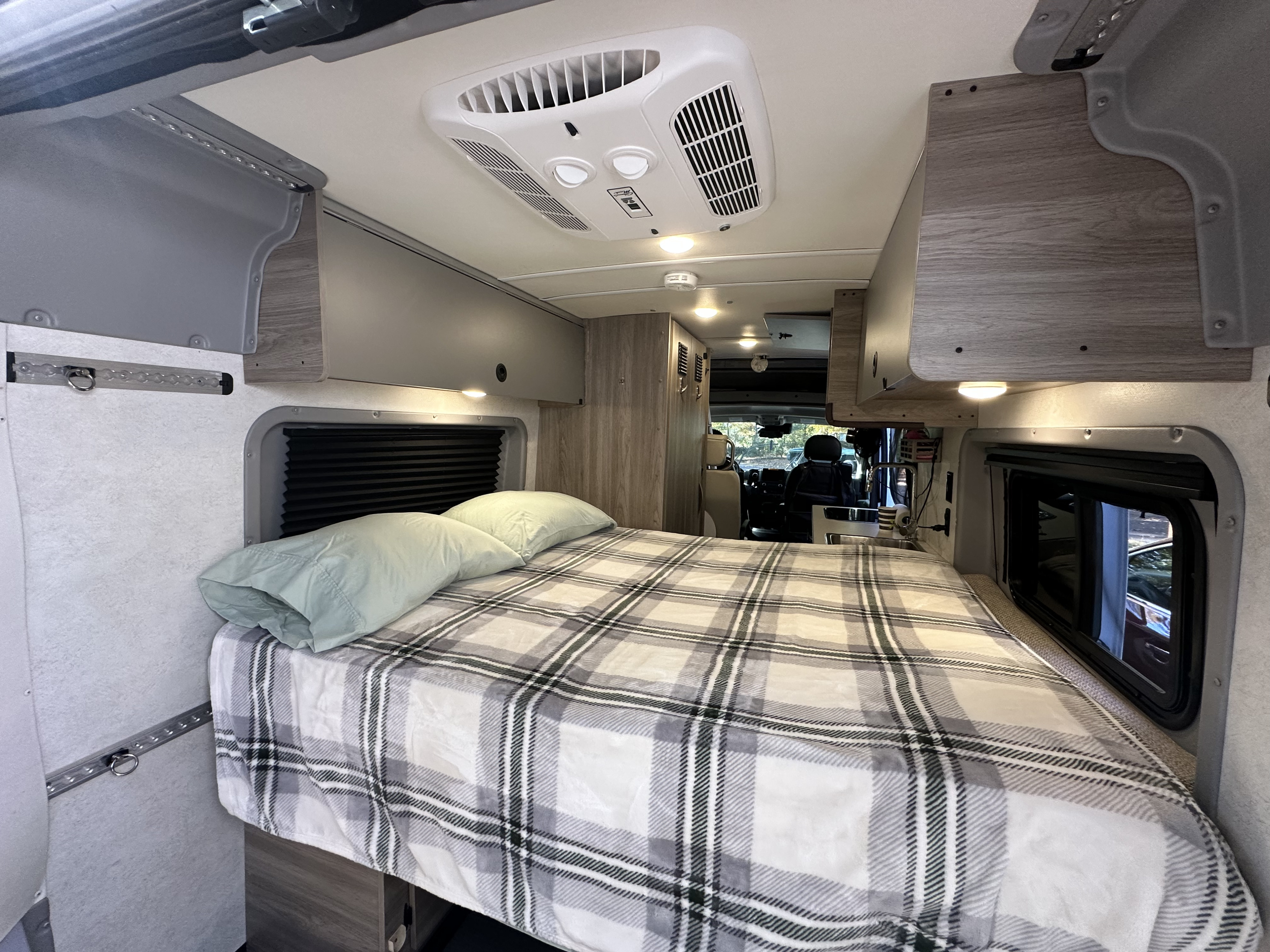 View of a modern RV rental interior featuring a bed with plaid bedding, overhead storage, and ceiling AC, extending to the kitchen.. Winnebago Solis 2023