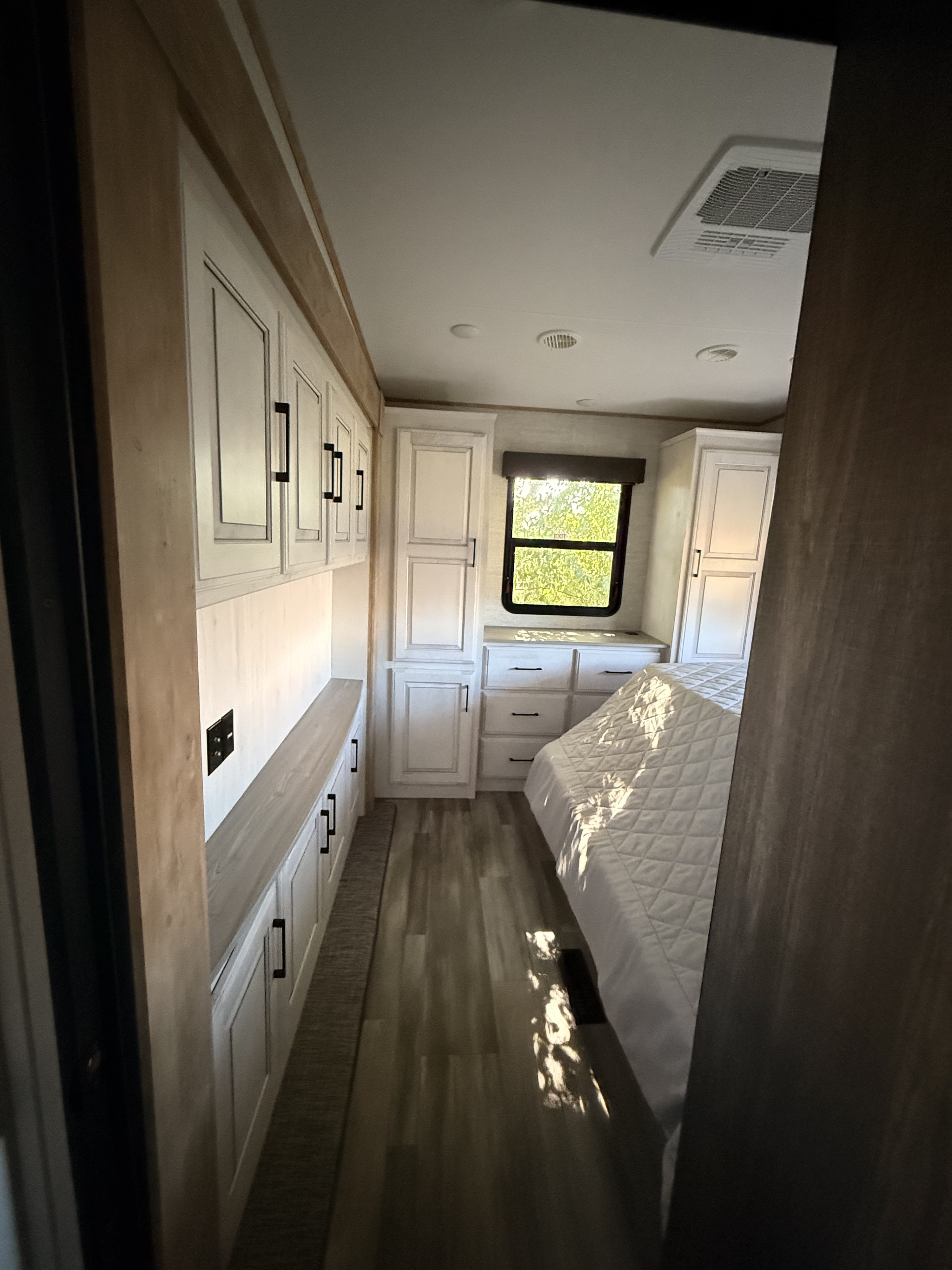 Bright RV rental bedroom with white cabinets, wood flooring, and a bed under a window showcasing green trees.. Heartland RVs Bighorn Traveler 2024