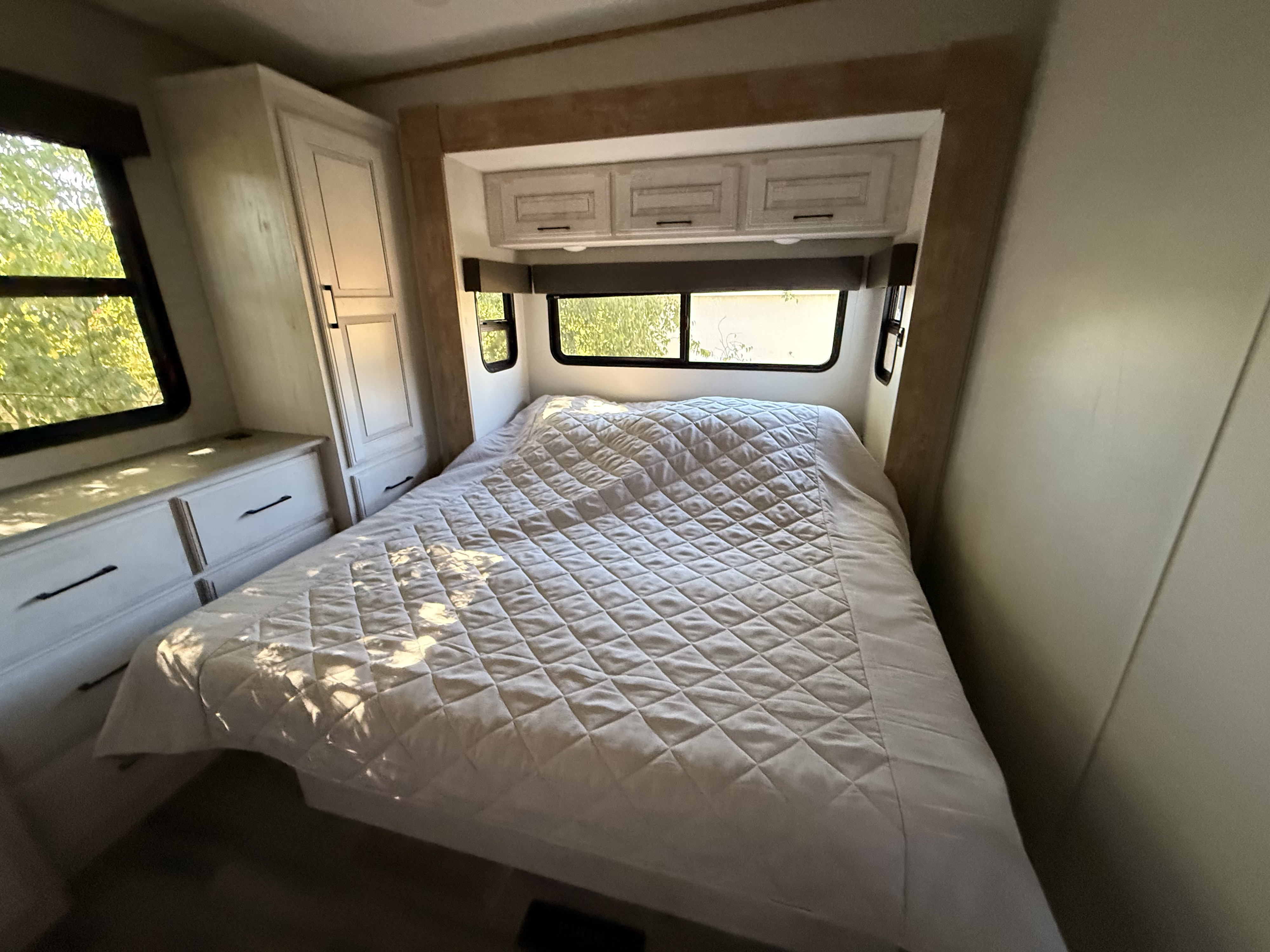 A cozy RV rental bedroom features a large bed with a light quilted bedspread, windows, and light wood cabinets.. Heartland RVs Bighorn Traveler 2024