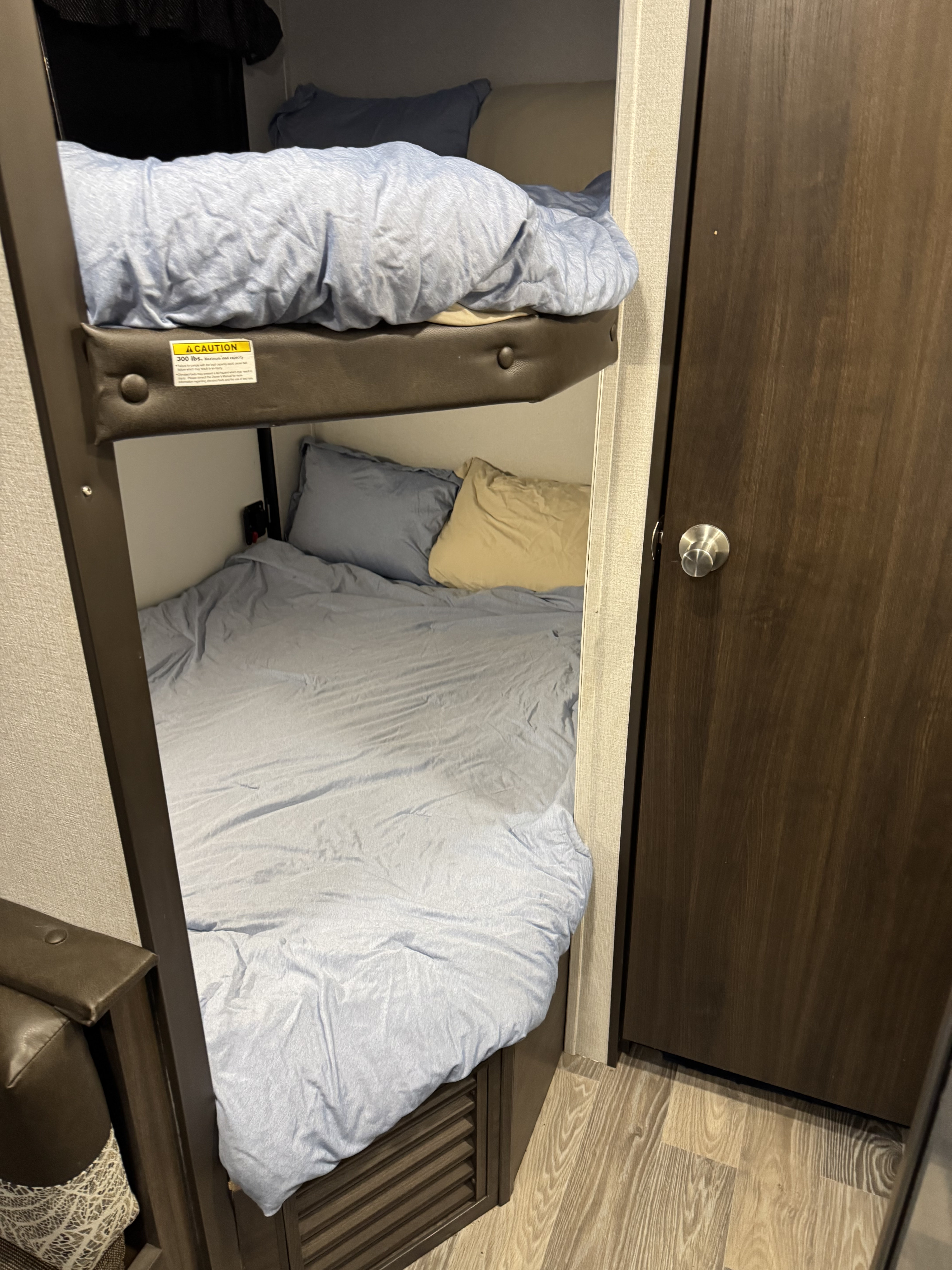 This RV rental features a cozy bunk bed sleeping area with blue bedding, a 300lb top bunk limit, and a nearby wooden door.. Keystone RV Springdale 2020