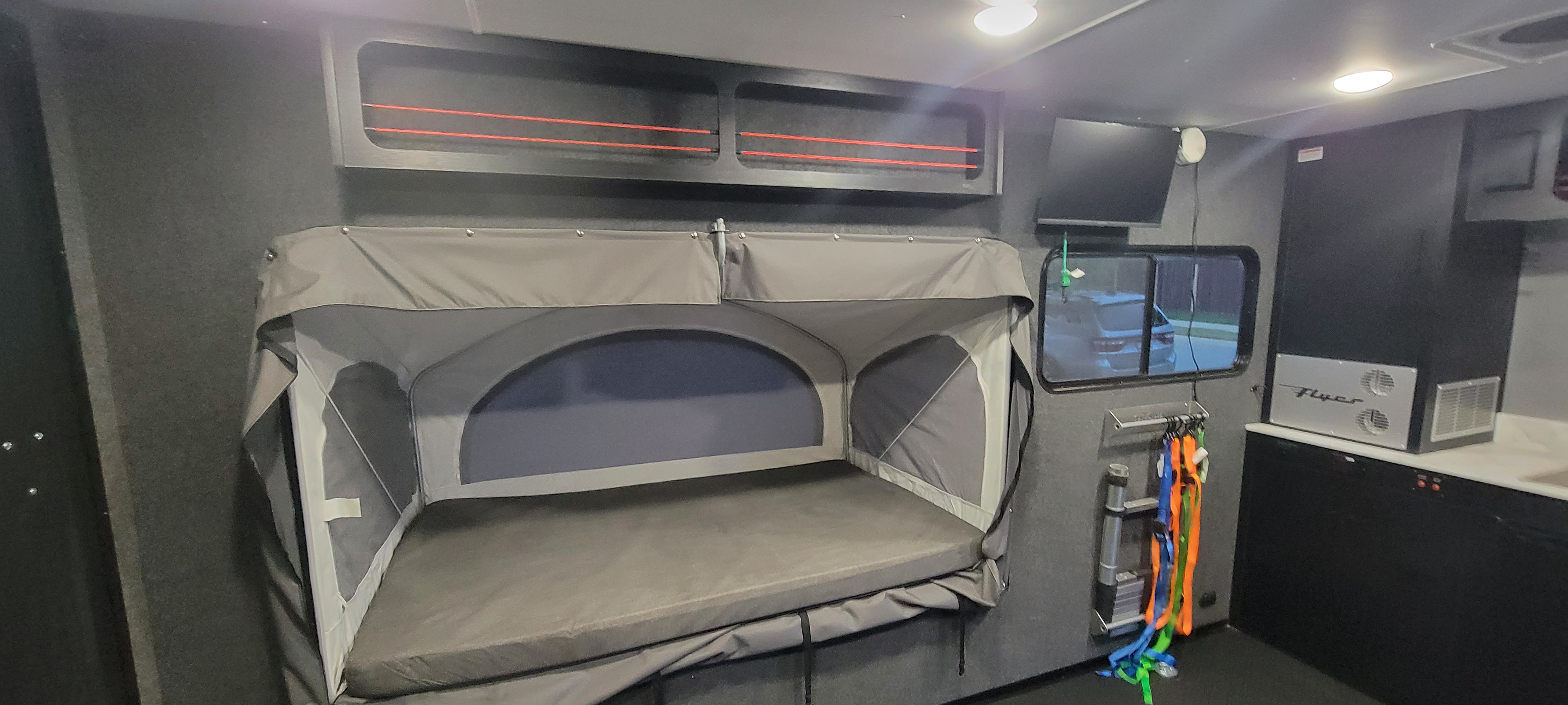 RV rental interior: grey fold-down bunk, overhead storage with red bungees, TV, and compact kitchen with 'Flyer' appliance.. InTech RV Flyer Discover 2024