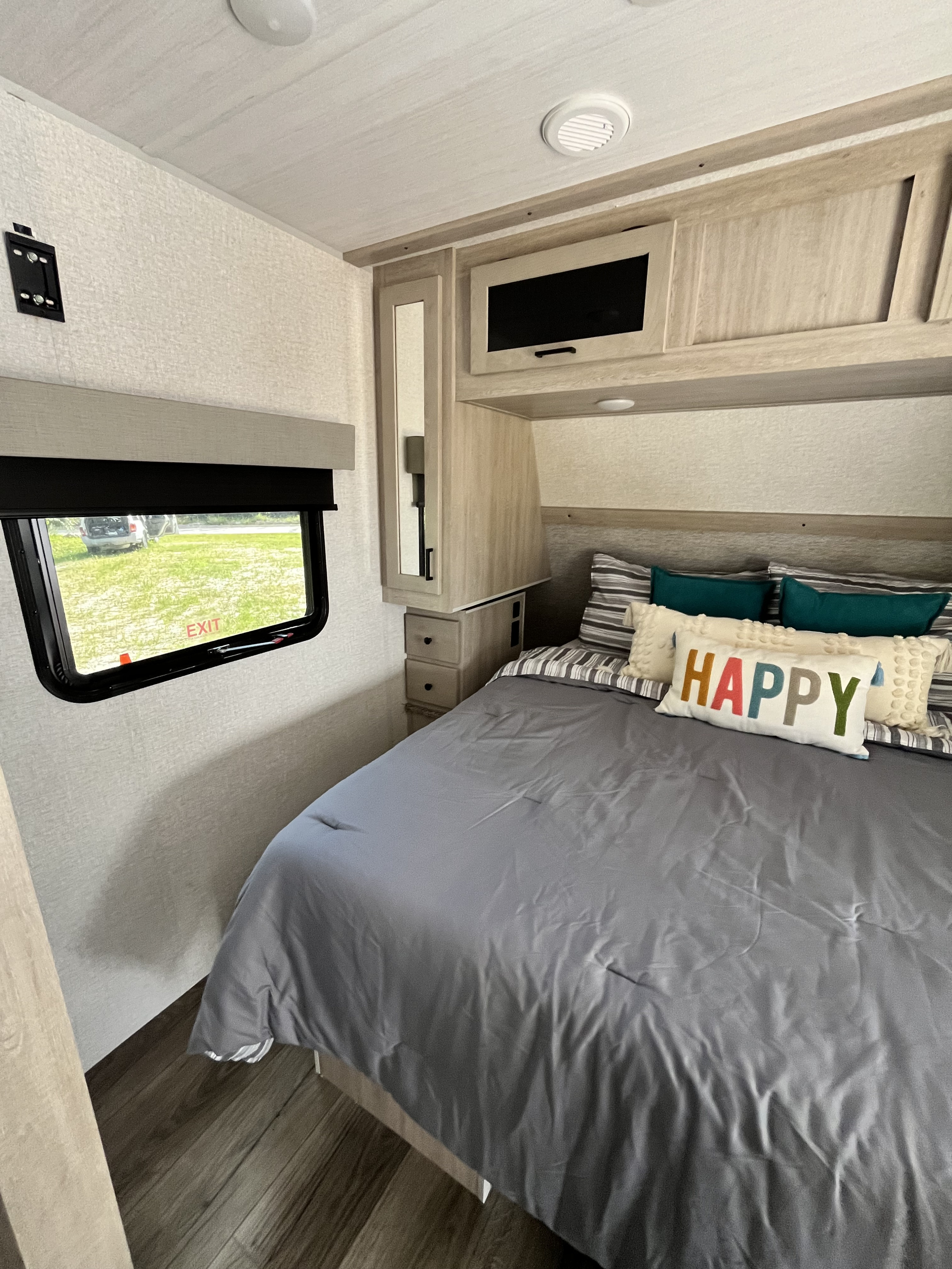 Cozy rv rental bedroom: bed with "HAPPY" pillow, overhead storage, mirror, and window with an 'EXIT' sign showing grass outside.. East to West Alta 2022